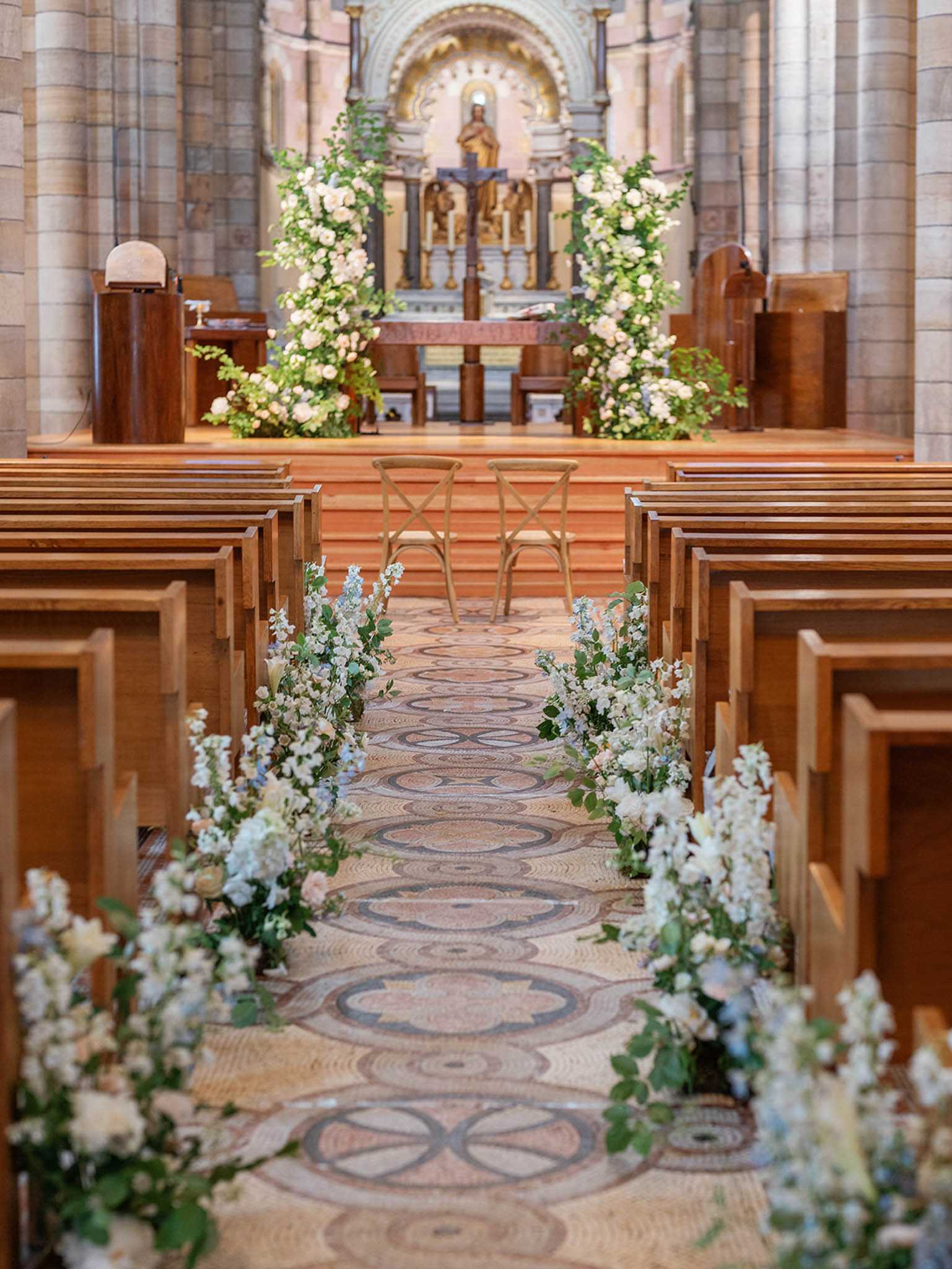 A pre-ceremony interior shot of a church set up for a wedding, with no guests or couple present. The aisle features a detailed geometric mosaic tile floor lined on both sides with loose floral arrangements placed directly on the ground, composed of white roses, white larkspur, pale blue delphinium, and green eucalyptus foliage in a garden-style arrangement. Two natural wood cross-back chairs are positioned at the altar step for the ceremony. At the altar, a large floral arch frames the crucifix and gilded altarpiece, constructed from white garden roses, greenery including trailing vines, creating a full, organic arch shape. The church interior has warm-toned stone columns and wooden pews on either side of the aisle. The overall floral palette is white, soft blue, and green, with a classic yet natural styling approach. Wide shot taken from the back of the aisle looking toward the altar. Potential venue feature image.