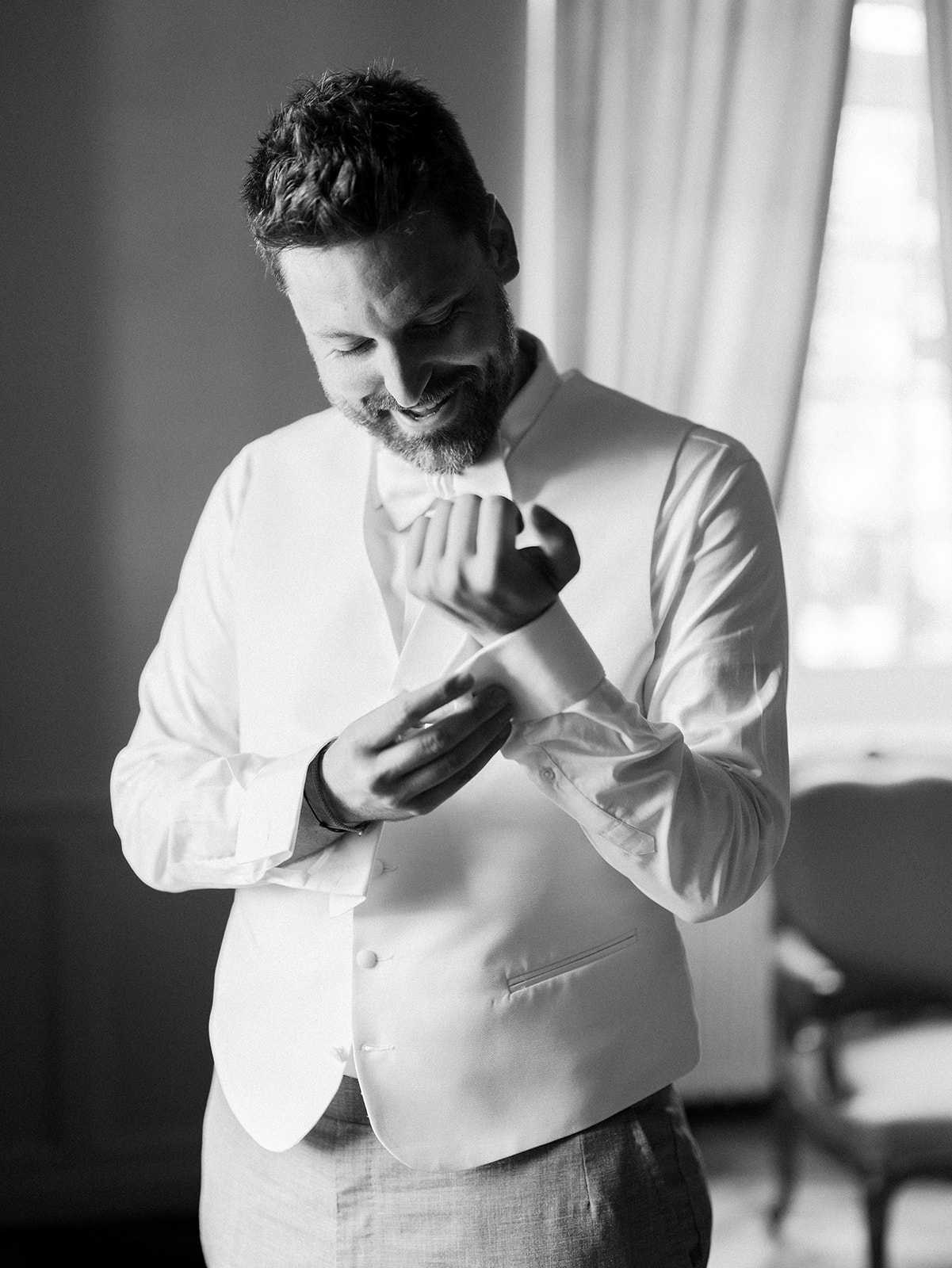 A black-and-white getting-ready portrait of the groom indoors, photographed against a bright window that provides soft backlighting with strong contrast between the light tones of his outfit and the darker background. He is smiling and looking down as he fastens his cufflinks, dressed in a white dress shirt, white waistcoat, and a bow tie, with dark trousers and a bracelet visible on his wrist. The image is a close-to-medium portrait shot with shallow depth of field, with a chair and curtains faintly visible in the background.