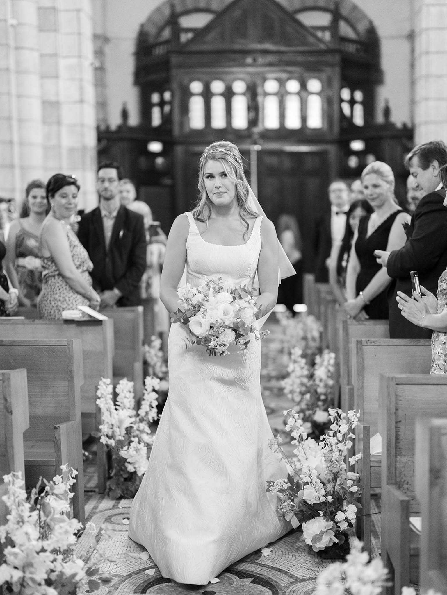 This black-and-white image captures the bride walking down the aisle during a church ceremony, shot from a medium portrait perspective at aisle level. The bride wears a structured square-neck, sleeveless gown with a textured fabric and a cathedral-length veil, and she is accessorized with a delicate floral or leaf headpiece; she carries a loose, garden-style bouquet of light-toned roses, ranunculus, and greenery with trailing eucalyptus. The aisle is lined on both sides with abundant low floral arrangements featuring tall stems — likely sweet peas, larkspur, and ranunculus — placed directly on the mosaic tile floor between wooden pews, creating a lush corridor effect. Approximately 20–30 guests are visible standing in the pews on either side, dressed formally in dark suits and evening wear, with a grand dark-wood organ or architectural screen visible in the background of the high-ceilinged church interior.