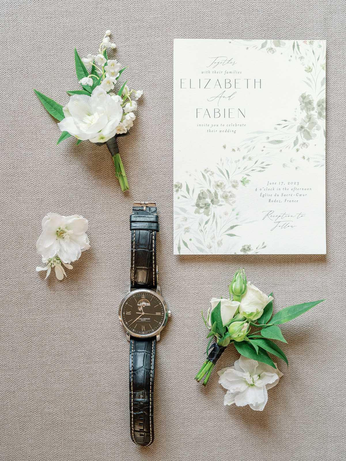 Flat lay detail shot of groom's wedding accessories arranged on a neutral linen-textured surface. The items include a wedding invitation card for Elizabeth and Fabien dated June 17, 2023 at Église du Sacré-Cœur in Rodez, France, featuring a soft watercolor botanical design in sage green and white; a Baume & Mercier dress watch with a dark dial and black crocodile-embossed leather strap; and three boutonnieres made with white ranunculus, lily of the valley, white spray roses with green buds, and fresh greenery. The color palette throughout is white, cream, and green, consistent with a classic garden-style wedding aesthetic.