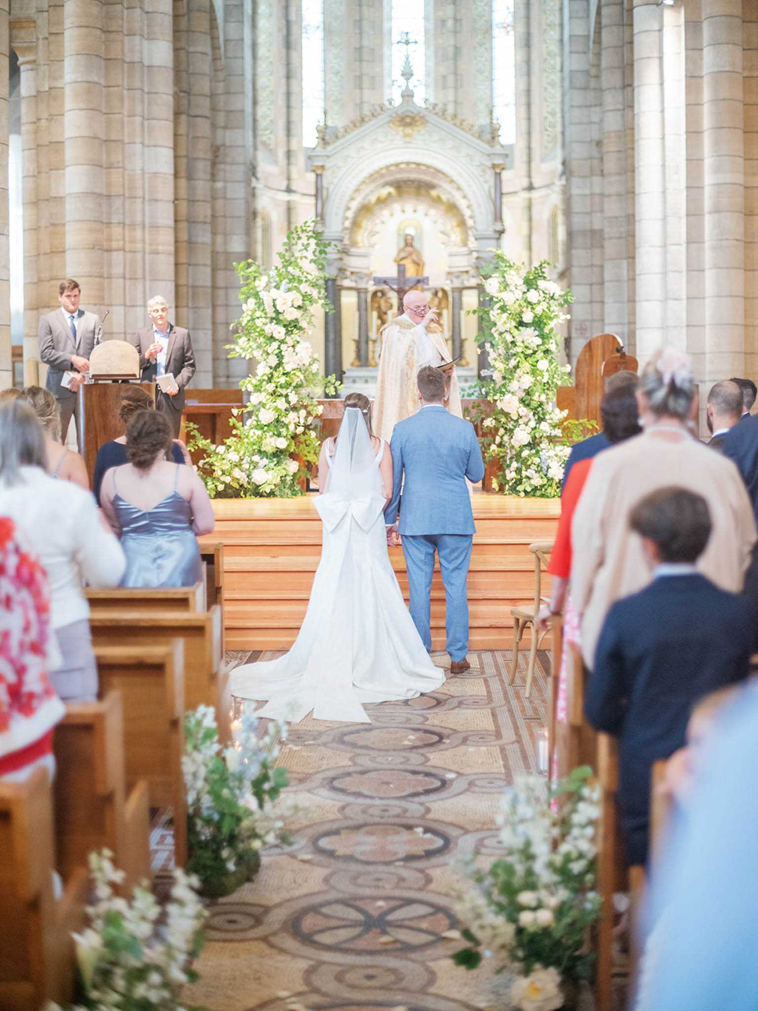 A wedding ceremony is taking place inside a large stone church with tall romanesque columns and an ornate altar. The bride, wearing a white gown with a long cathedral veil and a prominent bow detail at the back, stands alongside the groom in a light blue suit as a robed officiant reads from a book at the altar. Two large floral arrangements frame the altar, composed of white roses and abundant green foliage, while smaller clusters of white flowers and greenery line the mosaic-tiled aisle. Approximately 30 or more guests are seated in wooden pews on both sides, and a reader stands at a lectern to the left; the shot is taken from the back of the church looking toward the altar, giving a wide, central-aisle perspective.