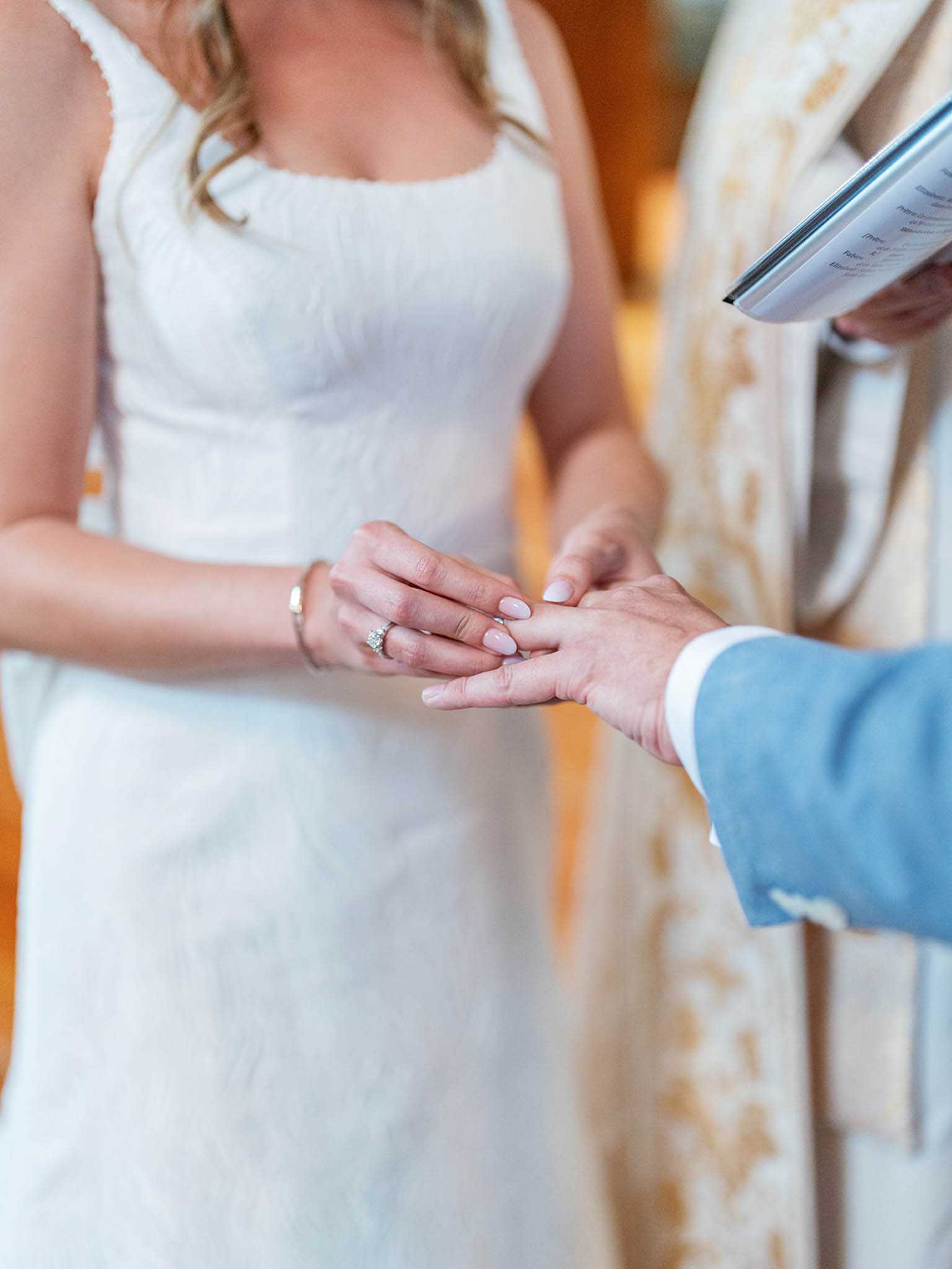 Close-up detail shot of a ring exchange during an indoor wedding ceremony. The bride, wearing a white square-neck fitted gown with subtle texture and a diamond engagement ring, places a wedding band onto the groom's finger; the groom is dressed in a light blue suit jacket. A officiant or priest is partially visible in the background wearing a white and gold ceremonial stole and holding an open ceremony booklet. The composition is tightly cropped on the couple's hands, keeping the background softly blurred.