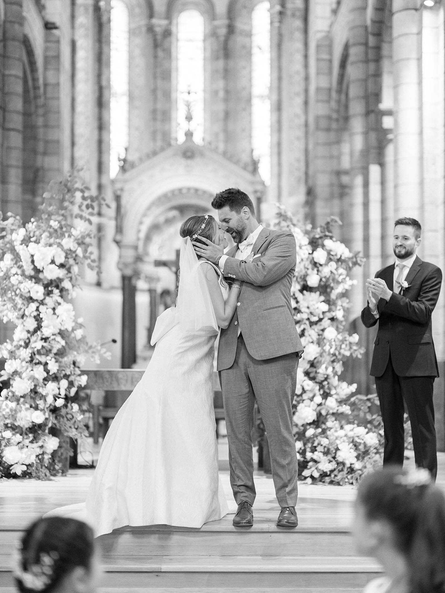 This black-and-white image captures the first kiss moment during an indoor religious ceremony held in a large stone church or cathedral with tall arched windows, ornate columns, and a decorative altar visible in the background. The bride wears a sleeveless fitted gown with a long veil and a floral hair accessory, while the groom is dressed in a light-toned suit with a bow tie; he cups her face with both hands as they kiss on a raised wooden platform. Two large freestanding floral arrangements frame the couple on either side, composed of full blooms — likely garden roses and ranunculus — with trailing foliage, appearing in bright whites and soft tones against the high-contrast B&W tones. A man in a dark suit, likely an officiant or best man, stands to the right applauding, and the heads of seated guests are visible in the foreground; the composition is a mid-range portrait shot taken from the congregation's perspective.