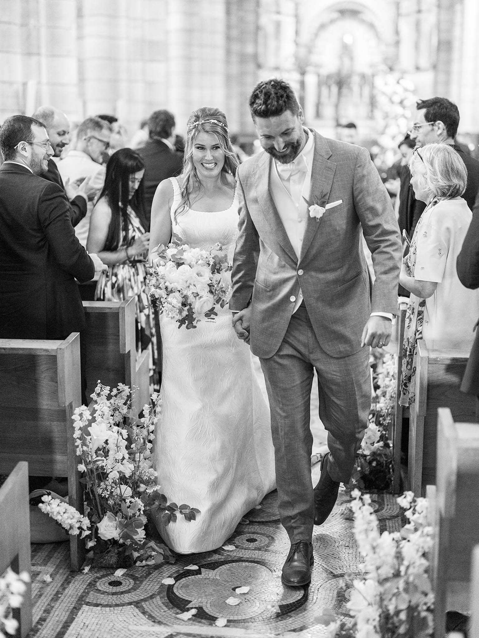 Black-and-white portrait of a bride and groom walking back down the aisle following their church ceremony, hand in hand and smiling. The setting is a grand stone church interior with arched architecture, ornate mosaic tile flooring, and wooden pews lining the aisle; scattered flower petals are visible on the floor. The bride wears a fitted sleeveless gown with a square neckline and a delicate floral headpiece, and carries a large loose bouquet of roses and foliage; the groom wears a light-toned suit with a boutonniere and a bow tie. Floral arrangements of delicate blooms — appearing to be sweet peas, roses, and ranunculus — are placed at the base of each pew along the aisle. Approximately 20–30 seated and standing guests are visible in the background. The image is shot in high-contrast black and white with bright highlights and deep shadows, captured as a mid-range portrait from the front.