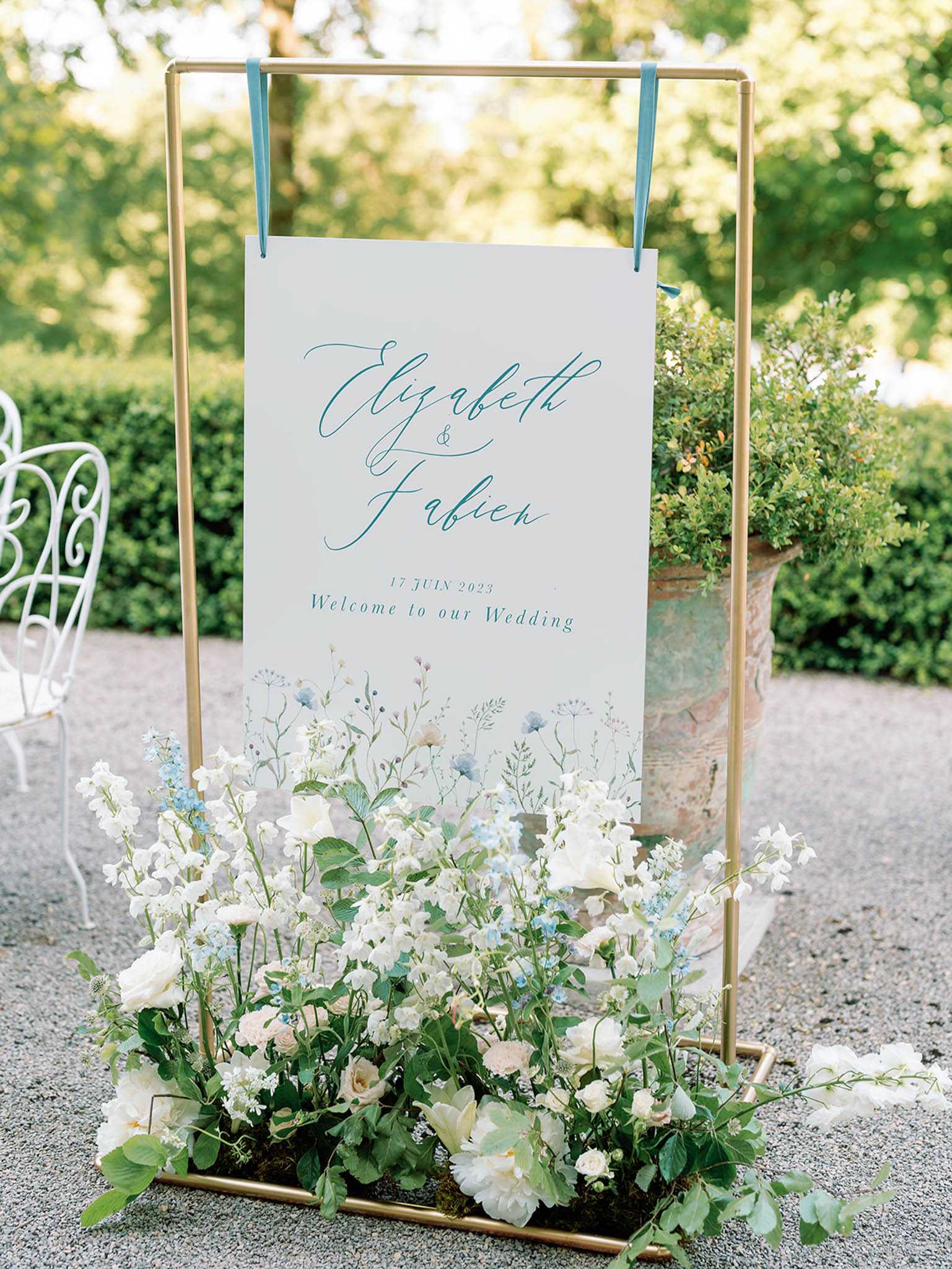 A close-up detail shot of an outdoor wedding welcome sign displayed on a slim gold metal frame stand, positioned on a gravel surface. The white sign features the names 'Elizabeth & Fabien' and the date '17 Juin 2023' in teal-blue calligraphy script, with 'Welcome to our Wedding' printed beneath, and a watercolor wildflower illustration along the bottom edge in soft blues, whites, and greens. The base of the gold frame is filled with a loose, garden-style floral arrangement of ivory ranunculus, white sweet peas, pale blush roses, blue forget-me-nots, and lush green foliage. A weathered terracotta urn with a topiary shrub is visible to the right, and a white wrought-iron chair appears in the background left, suggesting a classic French garden setting with a soft blue-and-white color palette throughout.
