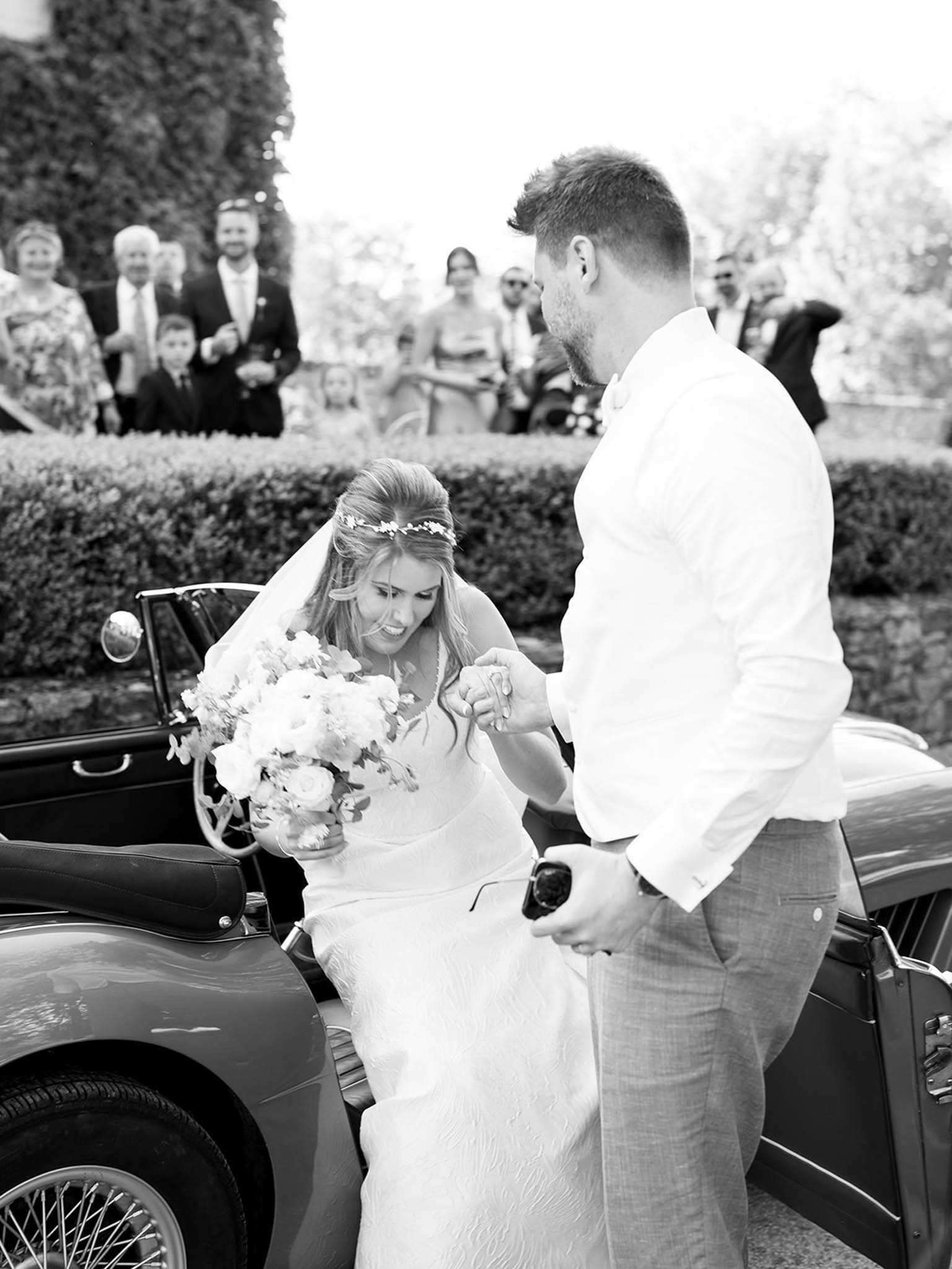 This black-and-white image captures the bride stepping out of a vintage convertible car, assisted by the groom who holds her hand. The bride wears a fitted white gown with a veil and a delicate floral hair piece, and carries a large, full bouquet of light-toned round blooms and trailing flowers. The groom is dressed in a white dress shirt and light-colored trousers. The scene is set outdoors, likely at a chateau or garden venue, with neatly trimmed hedgerows and an ivy-covered wall visible in the background. Approximately ten guests, including adults and children, watch the moment from behind a hedge line, some smiling. The image has strong contrast with bright highlights and deep shadows, shot at a mid-range distance capturing the full interaction between the couple.