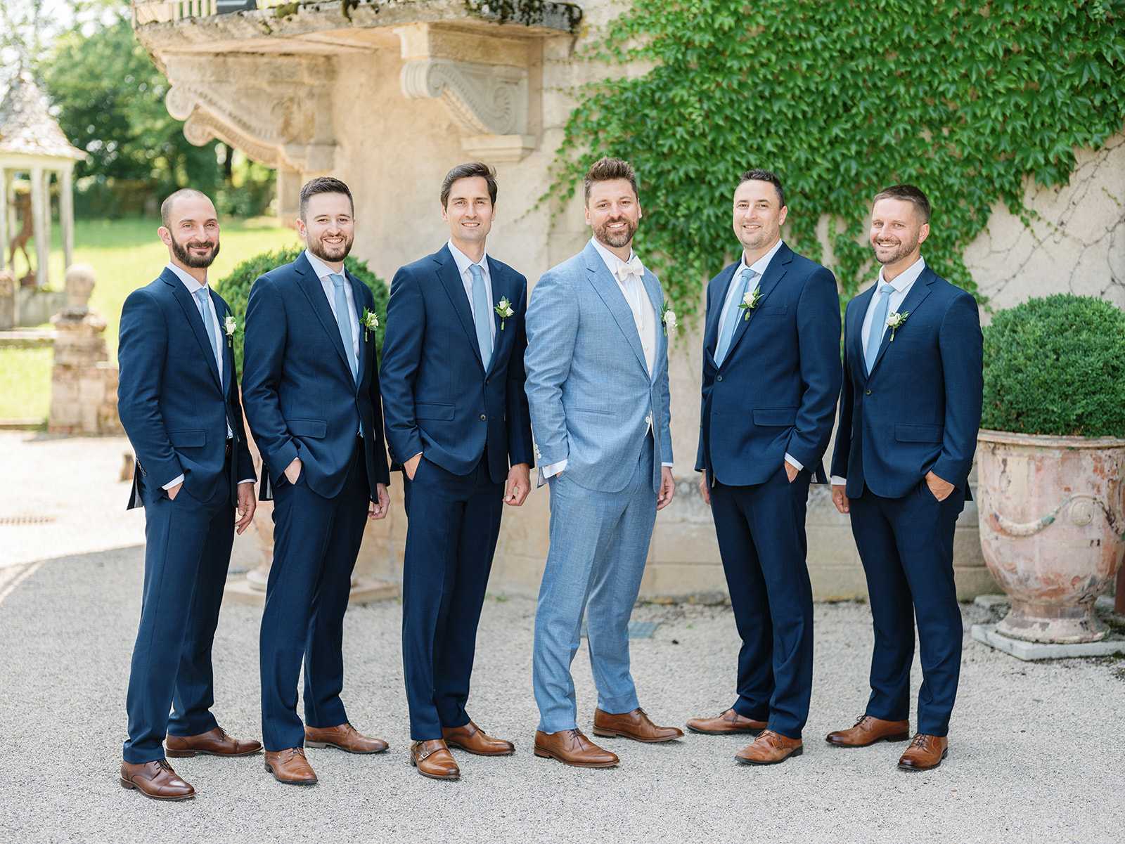 A groomsmen portrait taken outdoors on a gravel courtyard in front of a chateau facade with ivy-covered stone walls and ornate architectural details. Six men are posed in a loose group: the groom stands at center wearing a light blue suit with a white dress shirt and no tie, while his five groomsmen wear matching navy blue suits with light blue ties and tan leather oxford shoes. Each groomsman wears a white floral boutonniere with greenery, and the groom also has a matching boutonniere. The overall styling is classic with a blue tonal palette, and all men wear coordinating brown leather shoes. The shot is a mid-length group portrait taken at eye level in natural daylight.
