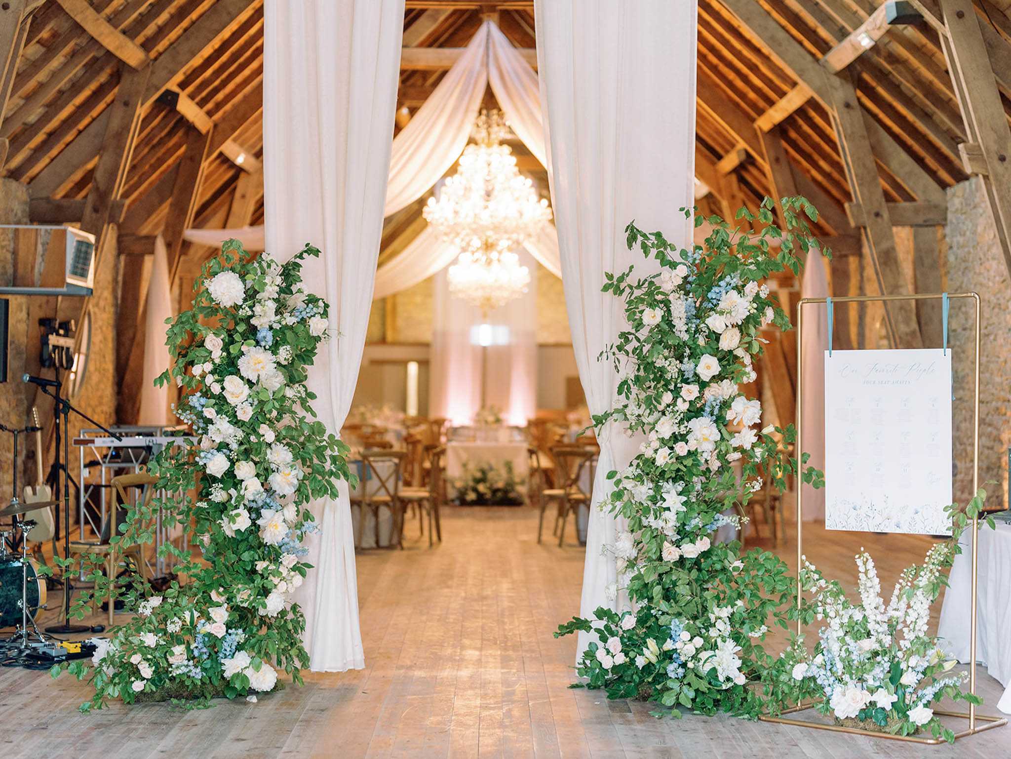 Wide shot of a wedding reception entrance inside a rustic barn venue with exposed wooden beam vaulted ceilings and stone walls. The entrance is framed by two tall floral columns featuring lush greenery, white peonies, white garden roses, soft blush roses, and powder blue delphinium, flanked by floor-length ivory fabric draping that extends up to the ceiling where a crystal chandelier hangs. To the right, a gold geometric frame holds a white seating chart sign with blue botanical illustrated borders, accompanied by a low floral arrangement at its base using the same white, blush, and dusty blue palette. The reception space beyond shows wooden cross-back chairs, draped tables with white linens, and warm uplighting in blush pink tones; a live band setup with instruments and speakers is visible on the left side. The overall decor palette is white, blush, dusty blue, and green with a garden-romantic aesthetic combining rustic barn architecture with refined floral design. Potential venue feature image.