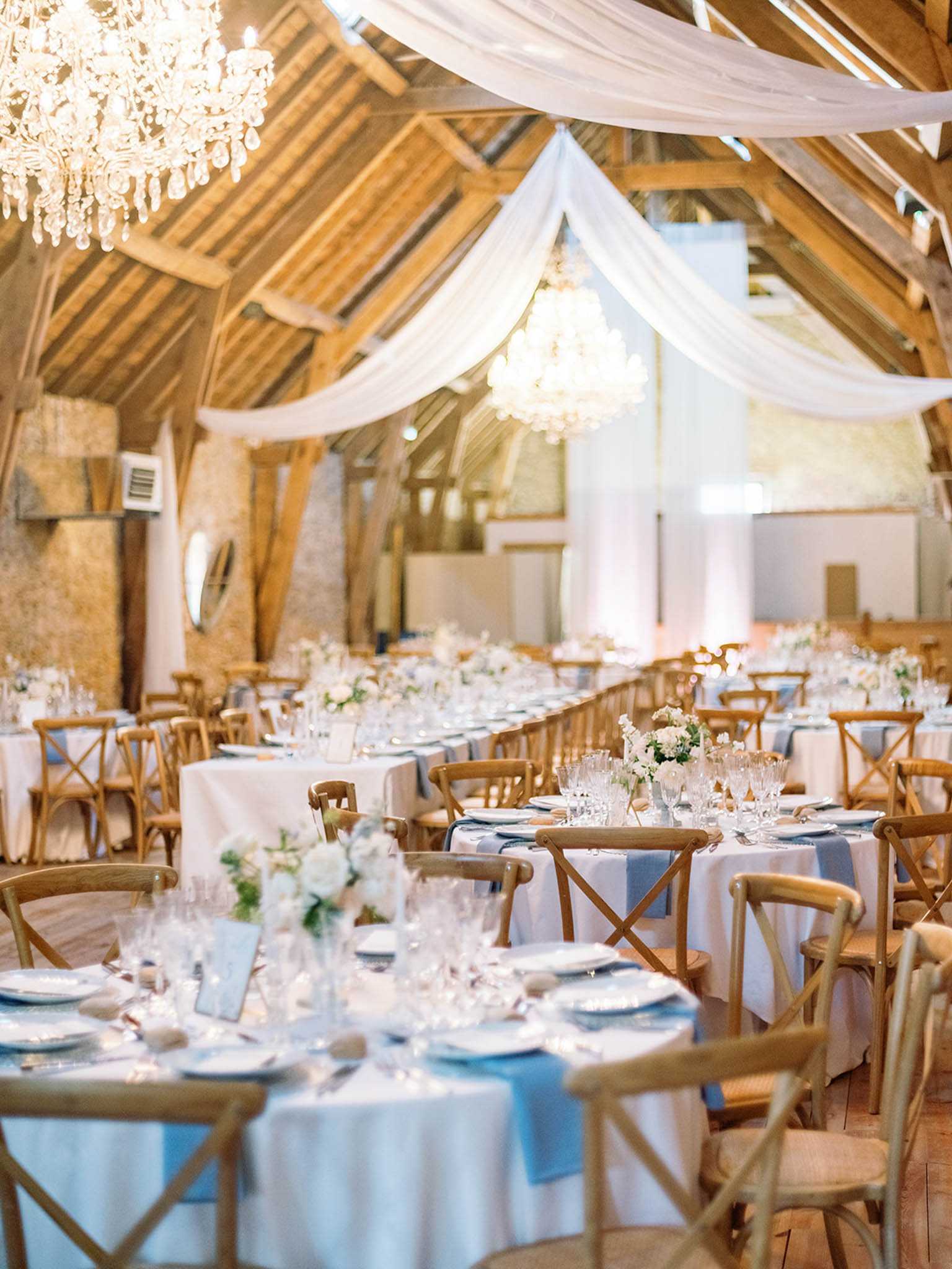 Indoor wedding reception setup inside a converted barn venue with exposed wooden beam vaulted ceilings and stone walls. The space is decorated with white fabric draping sweeping across the ceiling between two crystal chandeliers, creating a romantic, classic-rustic aesthetic. Round tables are dressed in white floor-length linens with dusty blue table runners, set with crystal glassware, white plates, and low centerpieces of white roses and greenery; natural wood cross-back chairs surround each table. The wide-angle shot captures the full length of the reception room, showing approximately eight to ten round tables fully set for a seated dinner with no guests present. Potential venue feature image.