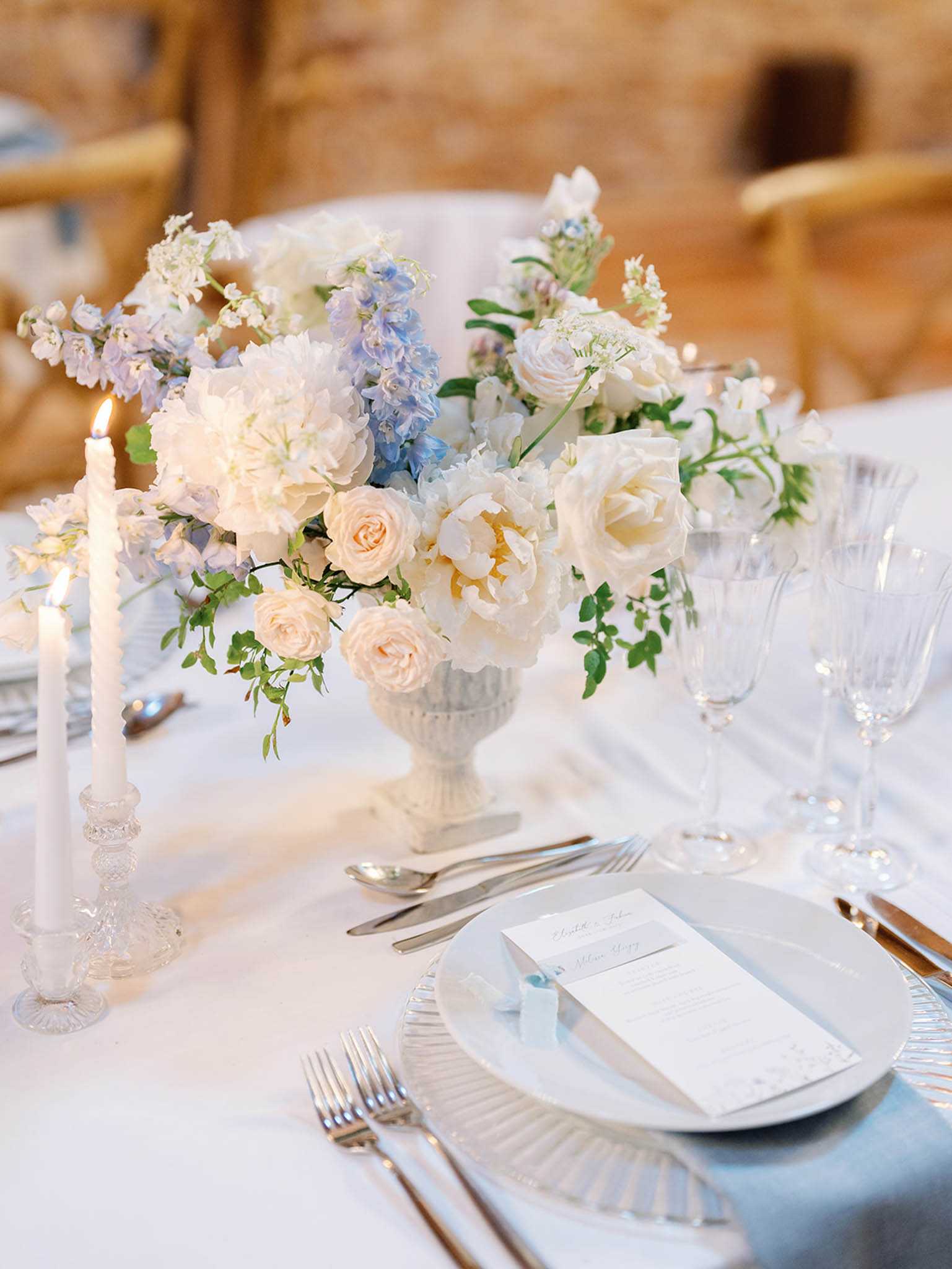Close-up detail shot of a wedding reception tablescape set in what appears to be a rustic indoor venue with warm stone walls visible in the background. The centerpiece features a white pedestal urn filled with blush garden roses, ivory peonies, pale blue delphiniums, white sweet peas, and trailing green foliage. Two white taper candles in a crystal candlestick holder are lit to the left of the arrangement. The place setting includes a ribbed glass charger plate layered with a white dinner plate, a light blue linen napkin, silver flatware, crystal glassware, and a folded white menu card tied with a pale blue ribbon. The overall decor palette is soft white, blush, and powder blue with a classic, refined styling.