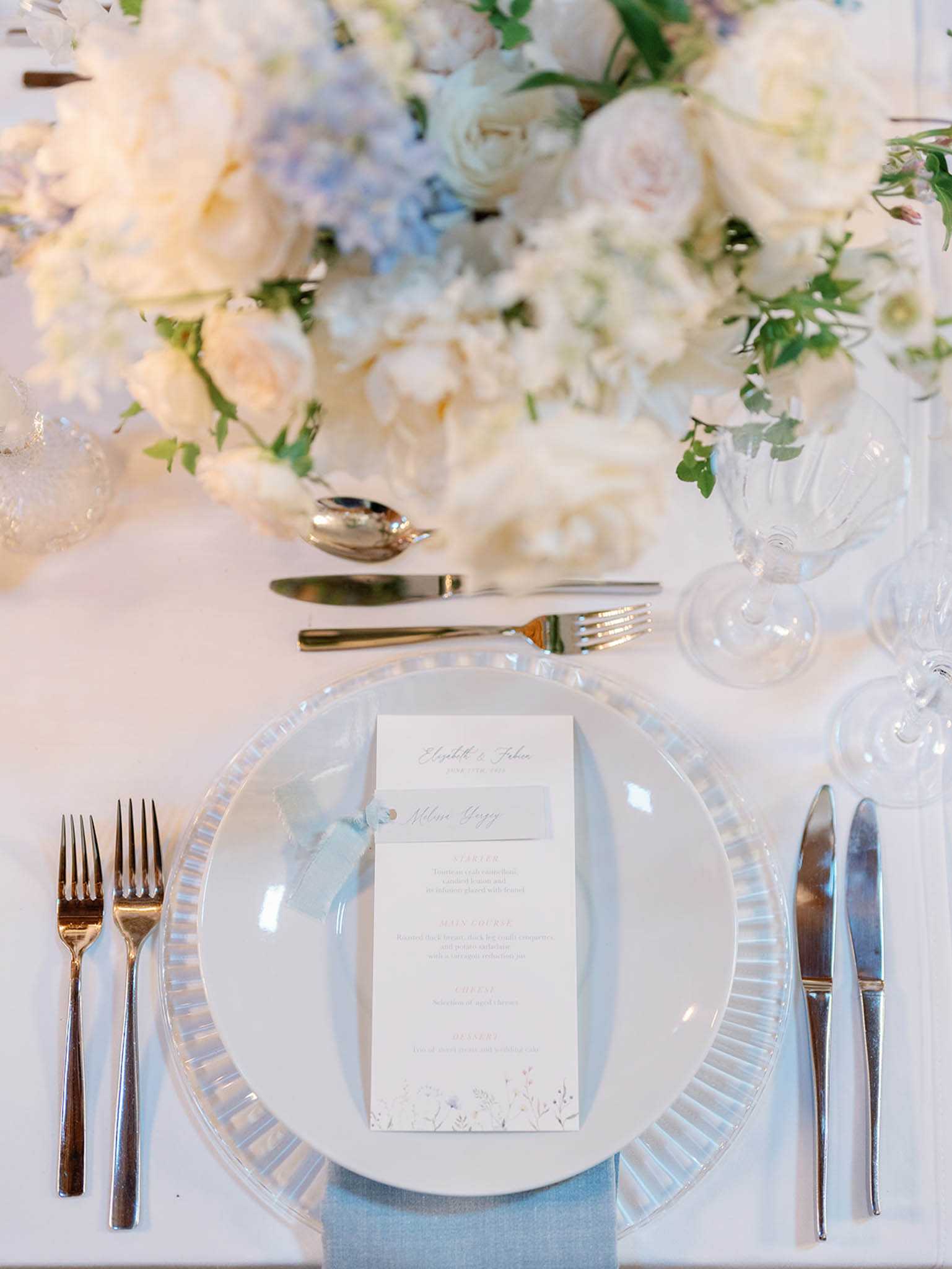 Close-up detail shot of a wedding reception table place setting styled in a soft blue and white palette. The place setting features a ribbed glass charger plate topped with a white dinner plate, a pale blue linen napkin, and a printed menu card with a wildflower botanical motif and a personalized name card attached with a light blue ribbon. Silver cutlery is arranged on both sides of the plate, with two forks to the left, a knife and butter knife to the right, and a spoon and additional fork placed horizontally above the plate. Two clear ribbed crystal glasses are positioned to the upper right. The background is filled with a voluminous low floral centerpiece composed of ivory garden roses, cream peonies, pale blue hydrangeas, white ranunculus, and trailing greenery, all on a white table linen.