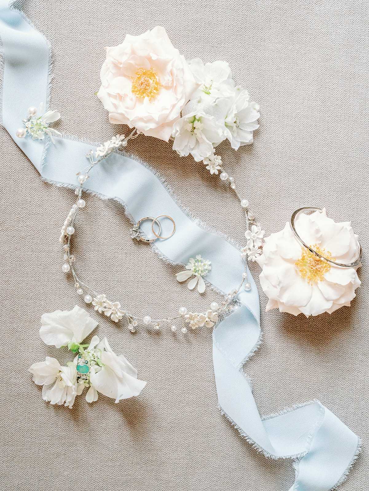 A flat lay of bridal accessories arranged on a neutral linen-textured surface, featuring a pale blue frayed silk ribbon winding through the composition. Two wedding bands and a diamond solitaire engagement ring rest on the ribbon at the center, surrounded by a delicate silver hair vine adorned with small freshwater pearls and white enamel floral details. Fresh blush and white peonies or garden roses are placed at the top and right, while white sweet peas and a small wrist corsage with a turquoise accent stone sit at the lower left. The overall color palette is soft and airy, combining pale blue, white, and blush tones in a romantic, classic bridal styling theme.
