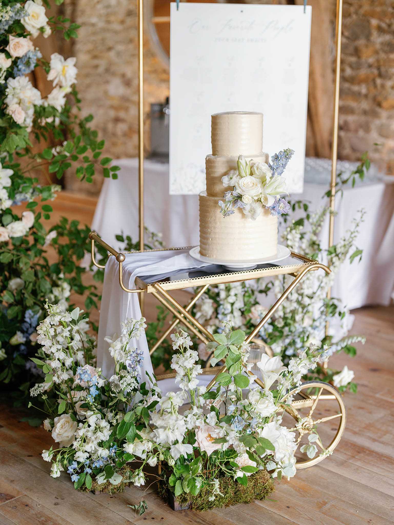 Detail shot of a three-tier wedding cake displayed on a gold bar cart inside what appears to be a barn venue with exposed stone walls and wooden beams. The cake has a textured buttercream finish in ivory-gold tones and is decorated with white roses, white lilies, and lavender-blue delphinium blooms. The gold cart is draped with soft grey-blue fabric and surrounded at the base by a lush ground-level floral arrangement of white sweet peas, white roses, pale blush roses, blue delphinium, and trailing greenery set on moss. A calligraphy wedding seating chart or welcome sign in mint green lettering is displayed on a gold frame behind the cake, and a large floral installation of blush roses and greenery is visible to the left. The overall decor palette combines ivory, soft blue, blush, and gold in a classic-romantic style.