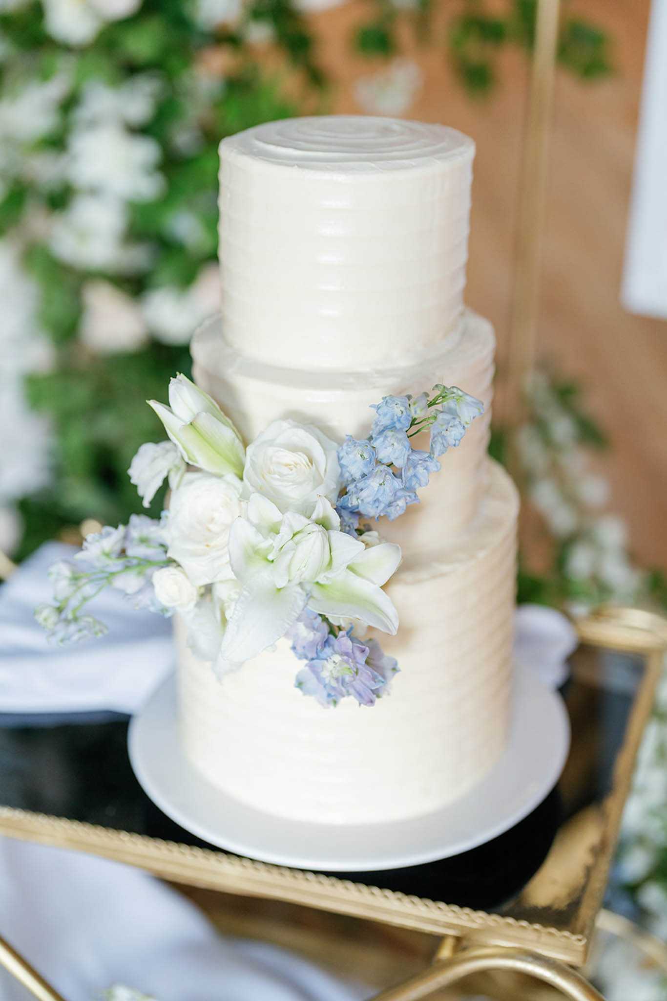 Close-up detail shot of a three-tier wedding cake with ivory buttercream frosting featuring a ribbed texture finish on each tier. The cake is decorated with a cascading floral arrangement of white ranunculus, white lilies, and periwinkle-blue delphinium blooms. It is displayed on a gold mirrored serving trolley with pale blue linen visible to the side, and a blurred background of green foliage and small white flowers. The overall decor palette combines ivory, soft blue, and gold tones in a classic style.
