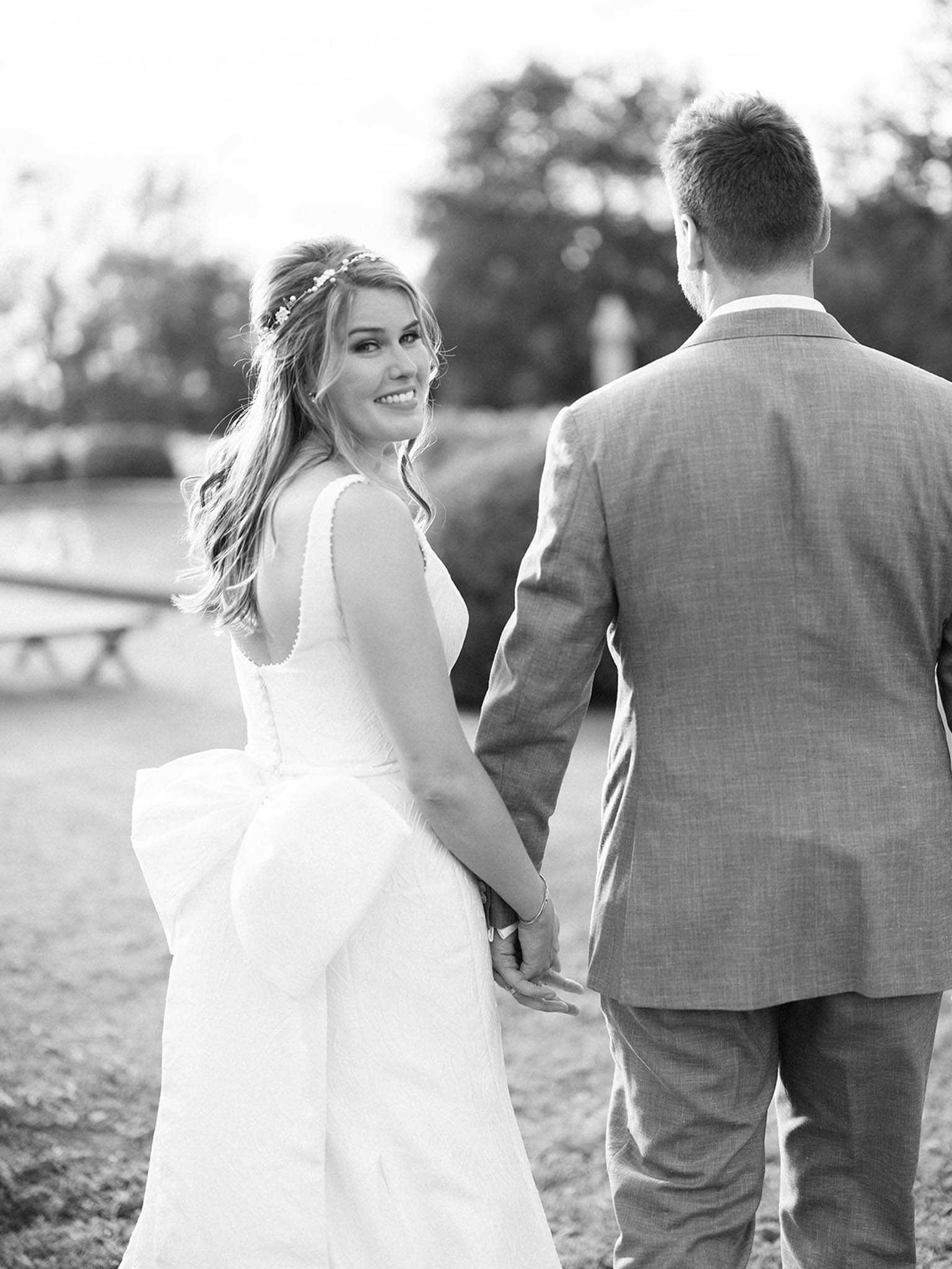 This is a black-and-white couple portrait taken outdoors, showing the bride and groom walking hand in hand while the bride turns back to look at the camera with a smile. The bride wears a sleeveless A-line gown with scalloped neckline detailing and a full skirt, and has loose wavy hair adorned with a delicate floral hair crown. The groom, whose back is fully turned to the camera, wears a light-toned structured suit jacket with a tie. The image is shot with soft, bright tones and a shallow depth of field, with a blurred outdoor setting visible in the background including what appears to be a bench or table. The composition is a medium portrait shot with gentle high-key lighting and smooth mid-tones throughout.