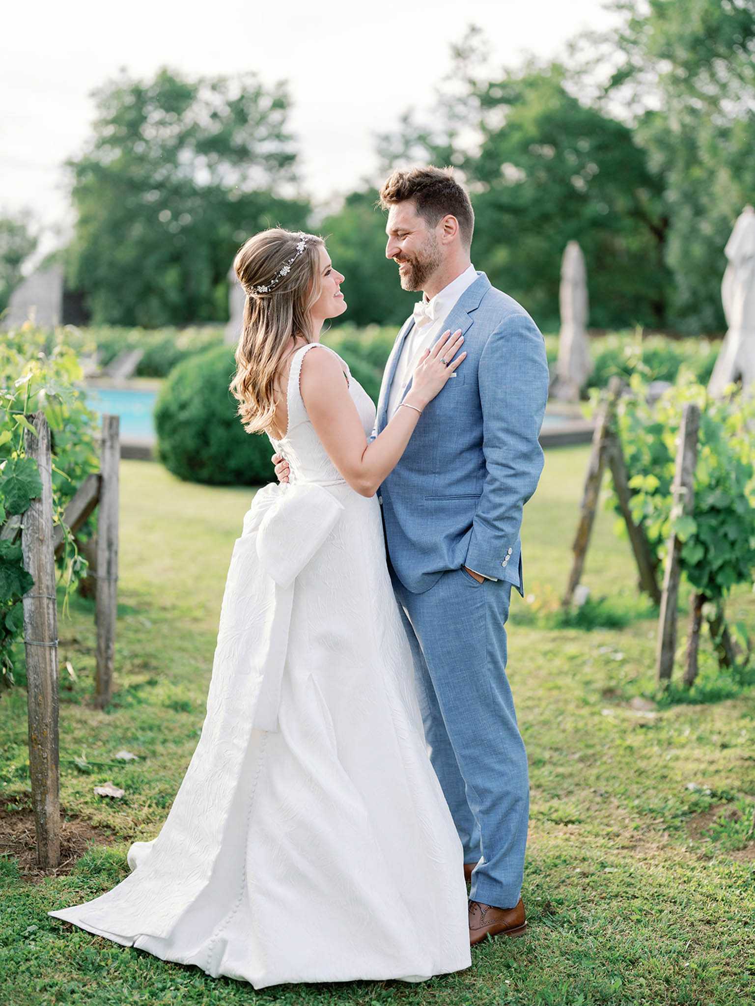 A couple portrait taken outdoors in a vineyard setting, with the bride and groom facing each other and smiling. The bride wears a white full-skirted ball gown with a large back bow detail, a low-cut neckline, and a delicate floral hair vine accessory worn with her hair half-up. The groom is dressed in a light steel-blue suit with a white dress shirt and no tie, paired with brown leather dress shoes. The shot is framed between rows of grapevines on wooden posts, with a pool and manicured topiary visible in the background. The composition is a mid-length portrait with soft, warm natural light typical of late afternoon.