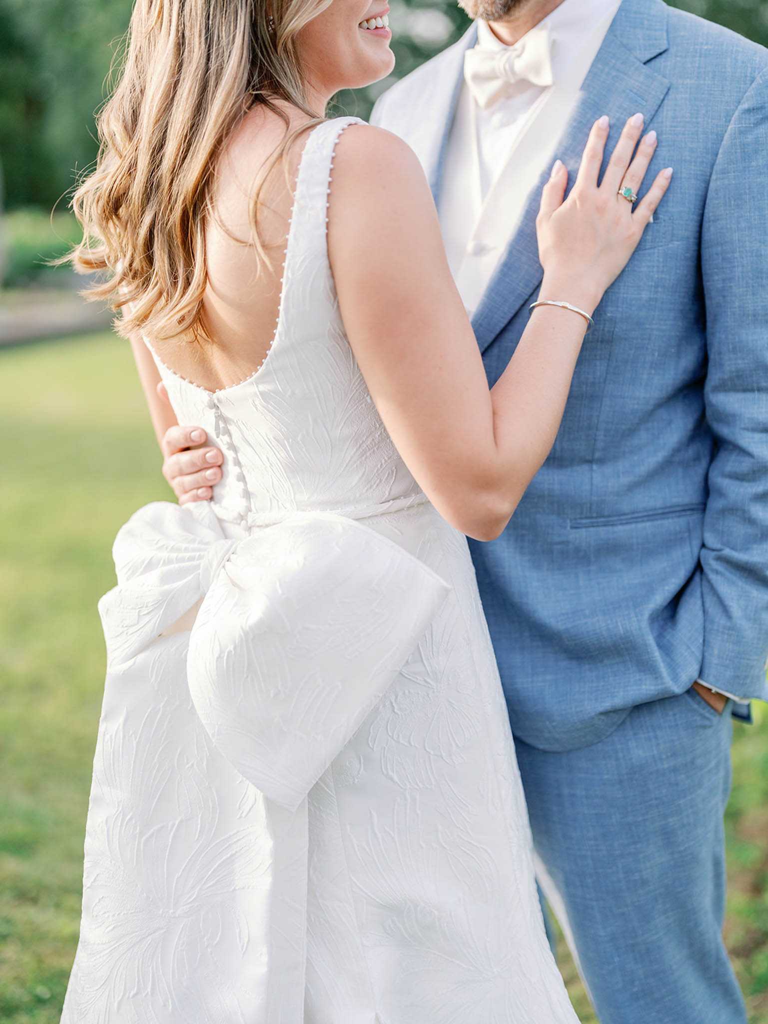 A close-up portrait of a bride and groom taken outdoors against a softly blurred garden background. The bride wears a white textured jacquard gown with a low scoop back, delicate pearl-trimmed straps, covered button closures, and a large structured oversized bow at the back waist; she accessorizes with a thin silver bracelet and an emerald-cut green stone ring. The groom wears a mid-blue linen suit with a cream bow tie and white dress shirt. The bride rests her hand on the groom's chest while he places his hand at her waist, and she is visibly smiling. The composition is a mid-detail crop focusing on the back of the dress and the couple's styling choices rather than their faces.