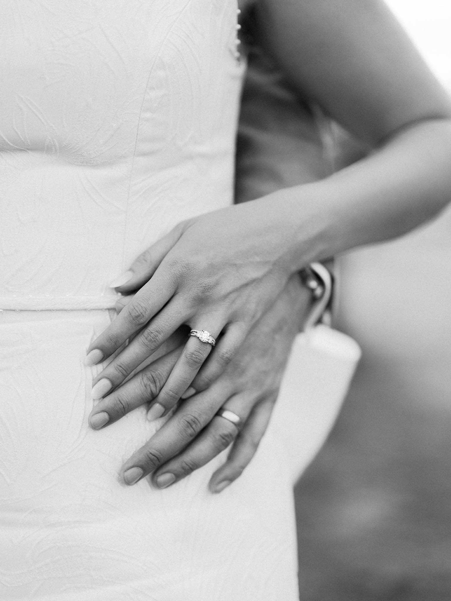 A black-and-white close-up detail shot of two sets of hands resting against the bride's wedding dress. The bride's hand displays a diamond solitaire engagement ring with a pavé band, while the groom's hand wears a plain band ring; a watch is partially visible on the groom's wrist. The bride's dress features a subtle textured or embroidered fabric pattern visible in the mid-section. The composition is tightly framed on the hands and waist, with soft contrast and light tones throughout, suggesting the couple is in an embrace.