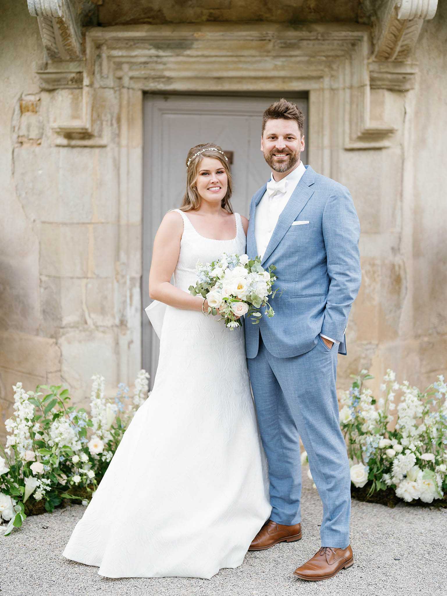 A couple portrait taken outdoors in front of an ornate stone doorway with a grey painted door, likely at a French chateau or historic estate. The bride wears a white sleeveless A-line gown with a square neckline and a delicate floral hair accessory, holding a loose bouquet of ivory garden roses, blush ranunculus, soft blue delphinium, and eucalyptus. The groom wears a light blue suit with a white bow tie and brown leather oxford shoes. Low floral arrangements of white and blush blooms with trailing greenery and touches of pale blue are placed at the base of the doorway on either side of the couple. The overall color palette is soft blue, white, and blush, giving the styling a classic, airy feel. Medium portrait shot, taken at eye level with a shallow depth of field.