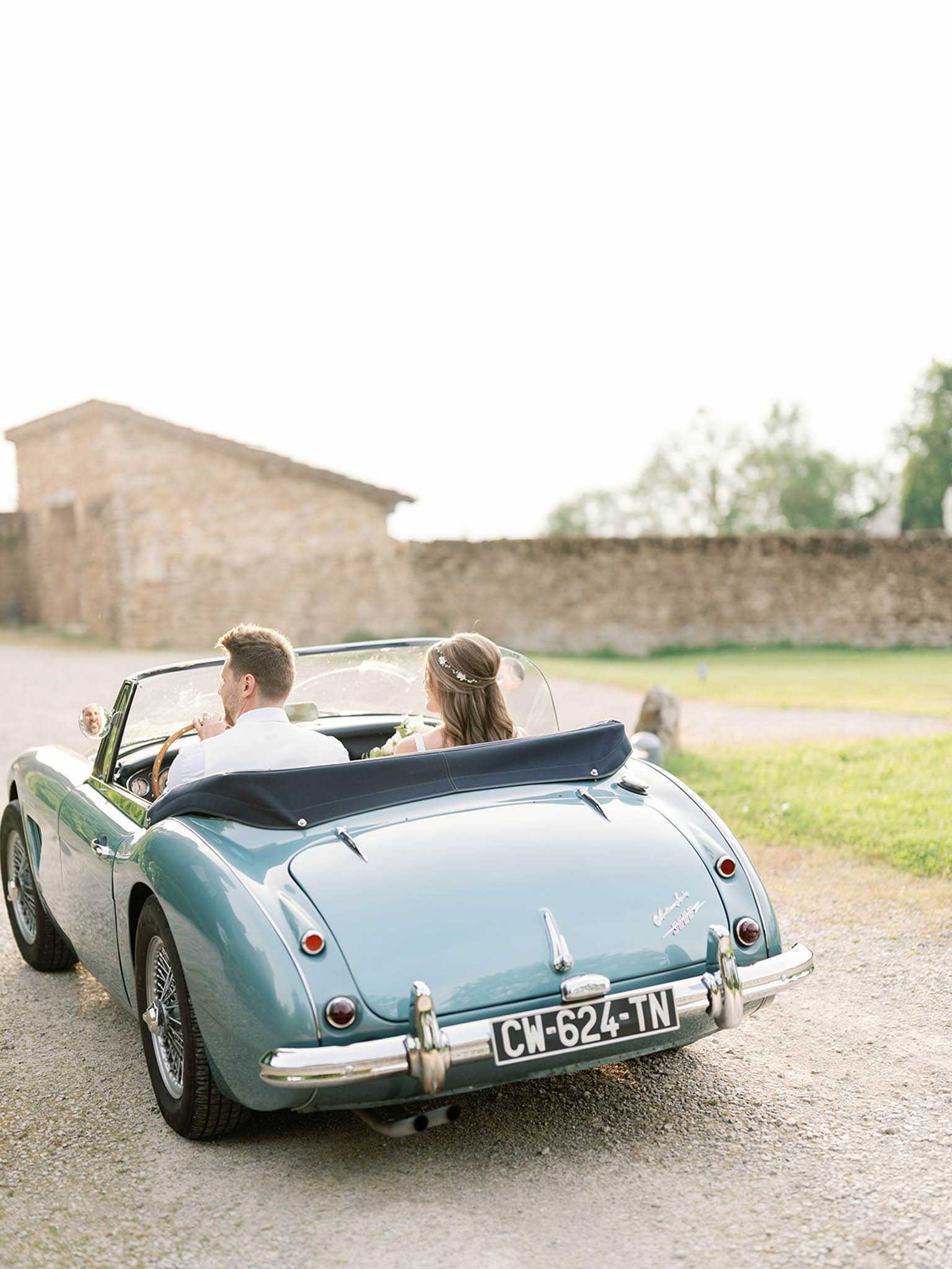 A bride and groom are seated in a vintage teal convertible sports car, shot from behind as they drive along a gravel path near a French stone building and low stone wall. The groom wears a white dress shirt and the bride has long brown hair adorned with a delicate floral or crystal hair vine. The car features chrome detailing, spoke wheels, a navy blue convertible top folded down, and a French license plate reading CW-624-TN. The composition is a wide rear shot capturing both the couple and the classic car as the central styling element, with soft warm ambient light suggesting late afternoon. The overall aesthetic is classic and romantic, with the vintage automobile serving as a deliberate styling prop.