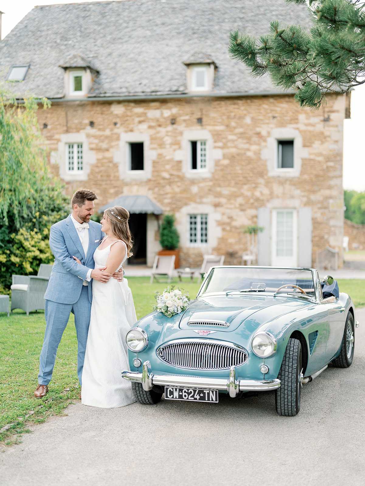 A couple portrait taken outdoors in front of a traditional French stone manor house, with the bride and groom standing beside a vintage teal convertible sports car (license plate CW-624-TN). The groom wears a light blue suit with a white bow tie and pocket square, while the bride wears a sleeveless white gown with a delicate headpiece and holds a loose bouquet of white blooms and greenery resting on the car's hood. The two face each other closely, with the groom's arms around the bride's waist. The vehicle is a classic open-top roadster in a teal-blue finish with chrome detailing, adding a retro styling element to the shoot. The composition is a medium-wide portrait with the stone building softly out of focus in the background. Potential venue feature image.