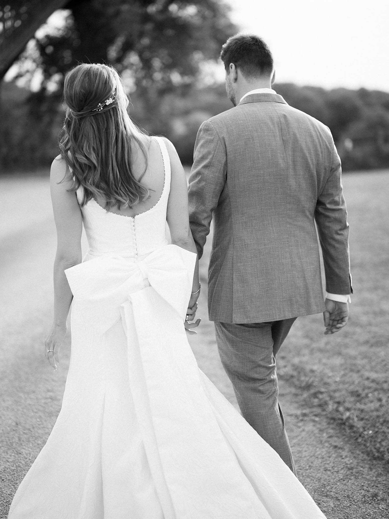 This black-and-white portrait shows a bride and groom walking away from the camera along a gravel path, holding hands. The bride wears a sleeveless ball gown with a prominent oversized bow at the back and a button-detailed bodice, with loose wavy hair accessorized by a delicate floral hair piece; the groom wears a light-toned structured suit. The image is shot from behind at a medium distance, with soft focus on the background, emphasizing the couple and the architectural detail of the gown's bow. High contrast between the bright white of the dress and the mid-tones of the suit gives the image a classic, editorial quality.