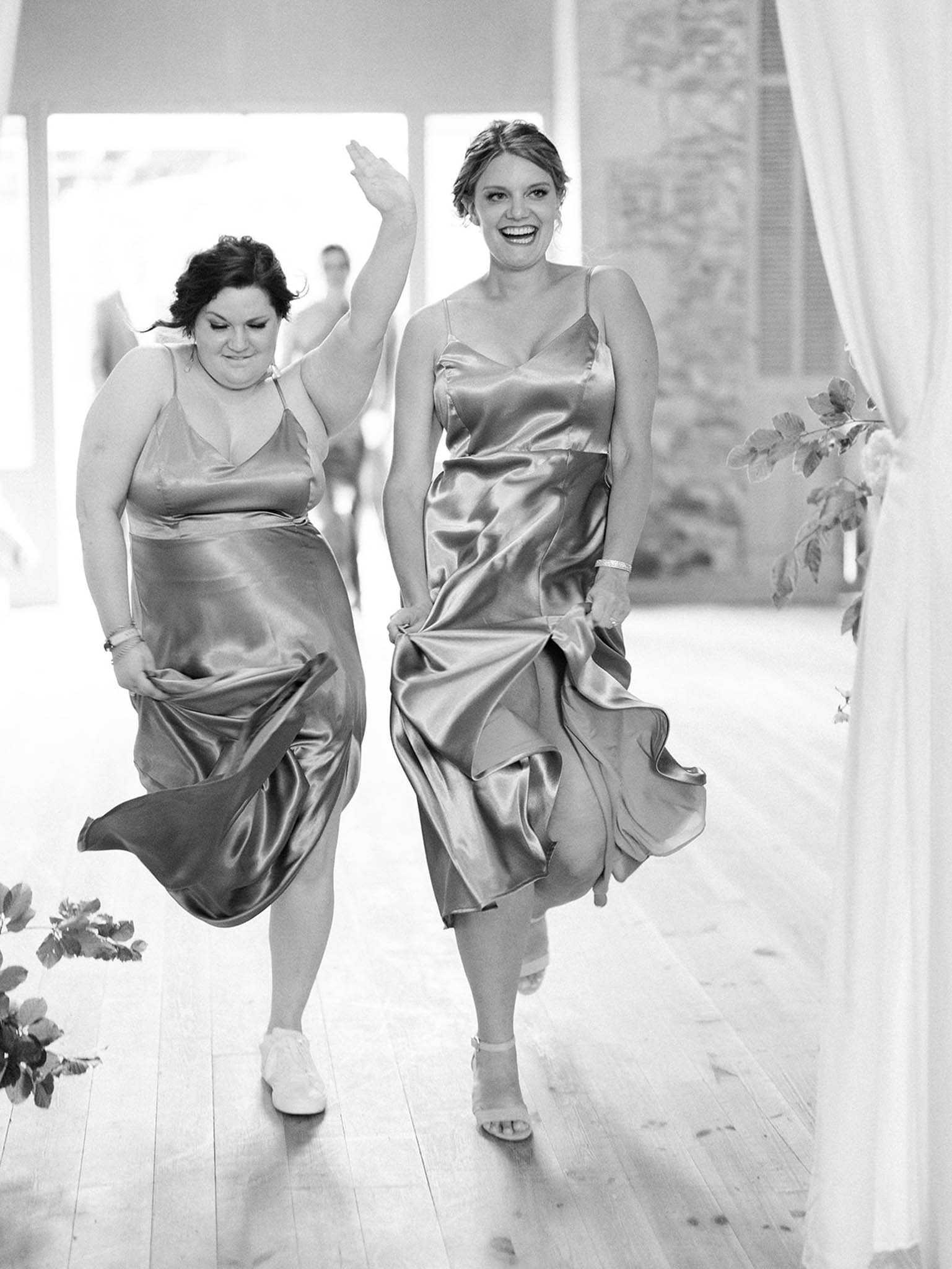 Black-and-white portrait-style shot of two bridesmaids making an energetic entrance into an indoor reception space, both lifting the hems of their satin slip-style midi dresses as they walk in stride. One woman is waving and laughing broadly while the other looks down with a playful expression; one wears white sneakers and the other open-toe heeled sandals. The room features light wood plank flooring, large windows letting in bright natural light, white draped fabric panels, and greenery accents visible at the edges of the frame. The high-contrast black-and-white tones highlight the sheen of the satin dresses and the movement of the fabric.