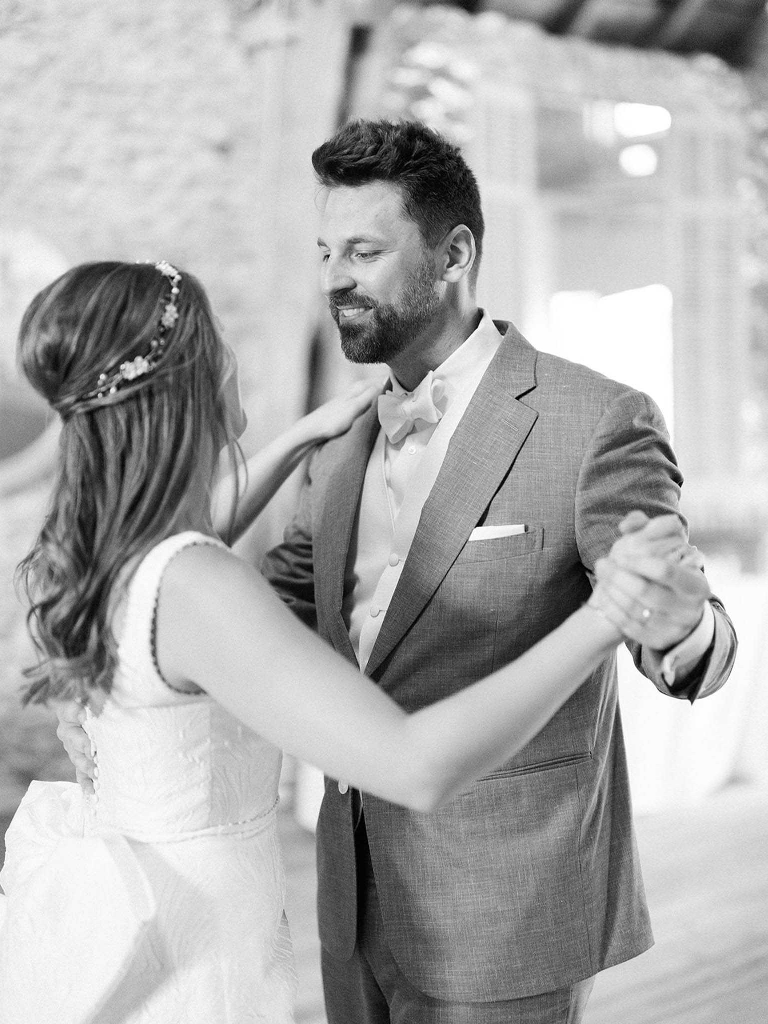 This black-and-white portrait captures a couple sharing their first dance, shot in a close-up style with a softly blurred background that suggests a rustic stone interior. The groom, who is smiling downward toward the bride, wears a textured suit jacket with a bow tie and a white pocket square; the bride, photographed from behind, wears a lace short-sleeve dress and an ornate floral hair vine woven through her long, loosely curled hair. The image has bright, airy tones with gentle contrast, giving it a soft film-like quality. The composition is an intimate mid-shot portrait focused on the couple's interaction and styling details.