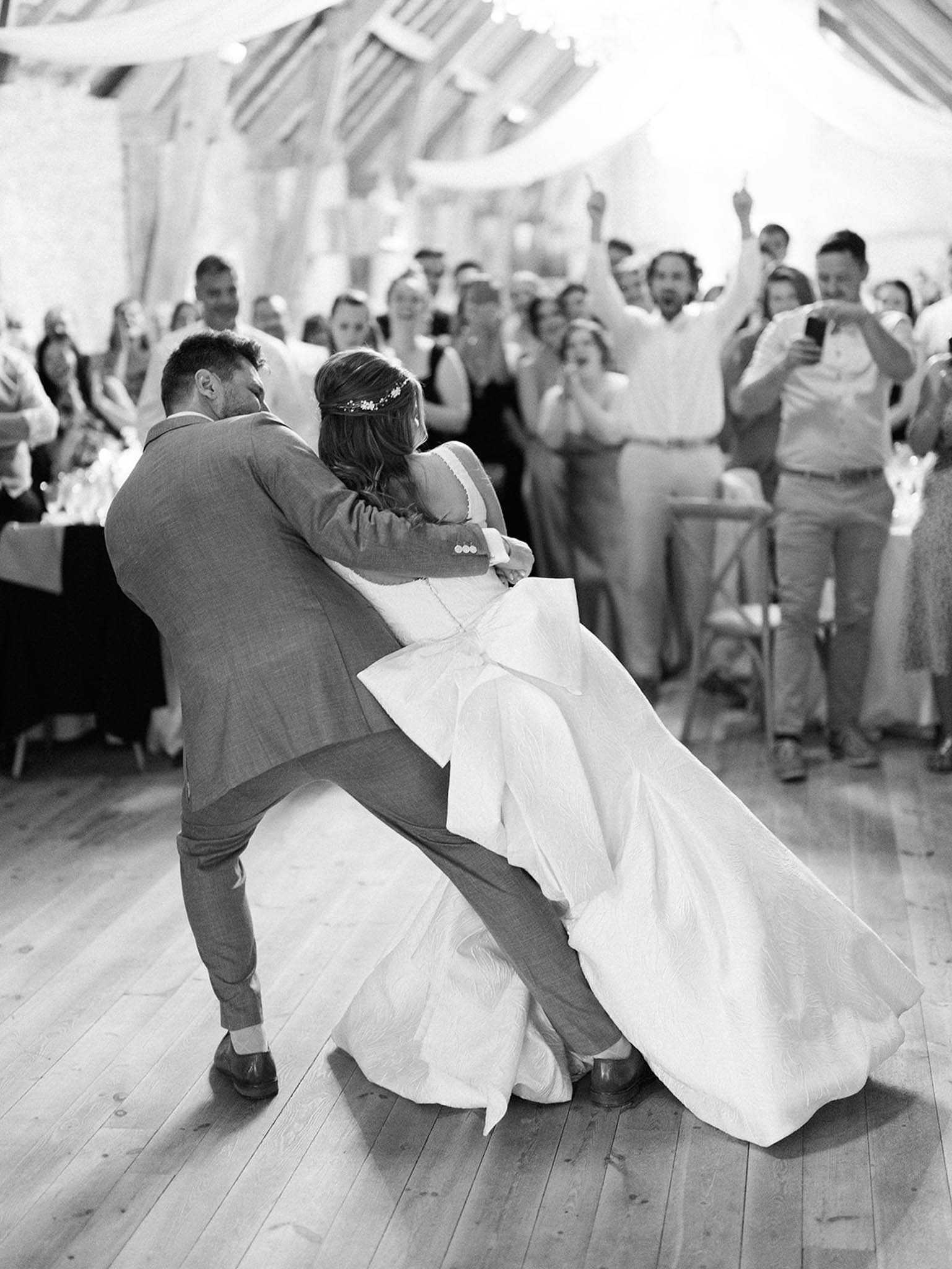 Black-and-white image of the couple's first dance inside a barn or marquee reception venue with exposed wooden roof beams and draped fabric overhead. The groom, wearing a suit, is dipping the bride low toward the wooden plank floor while she wears a full-skirted gown with a large back bow detail and a floral hair accessory. The dynamic dip moment is met with an enthusiastic reaction from a crowd of approximately 30–40 guests lining the dance floor behind them, several of whom have their arms raised and phones out. The image is shot from behind the couple at floor level, giving a wide portrait composition with strong contrast between the bright white dress and the mid-toned suit against the pale wooden floor.