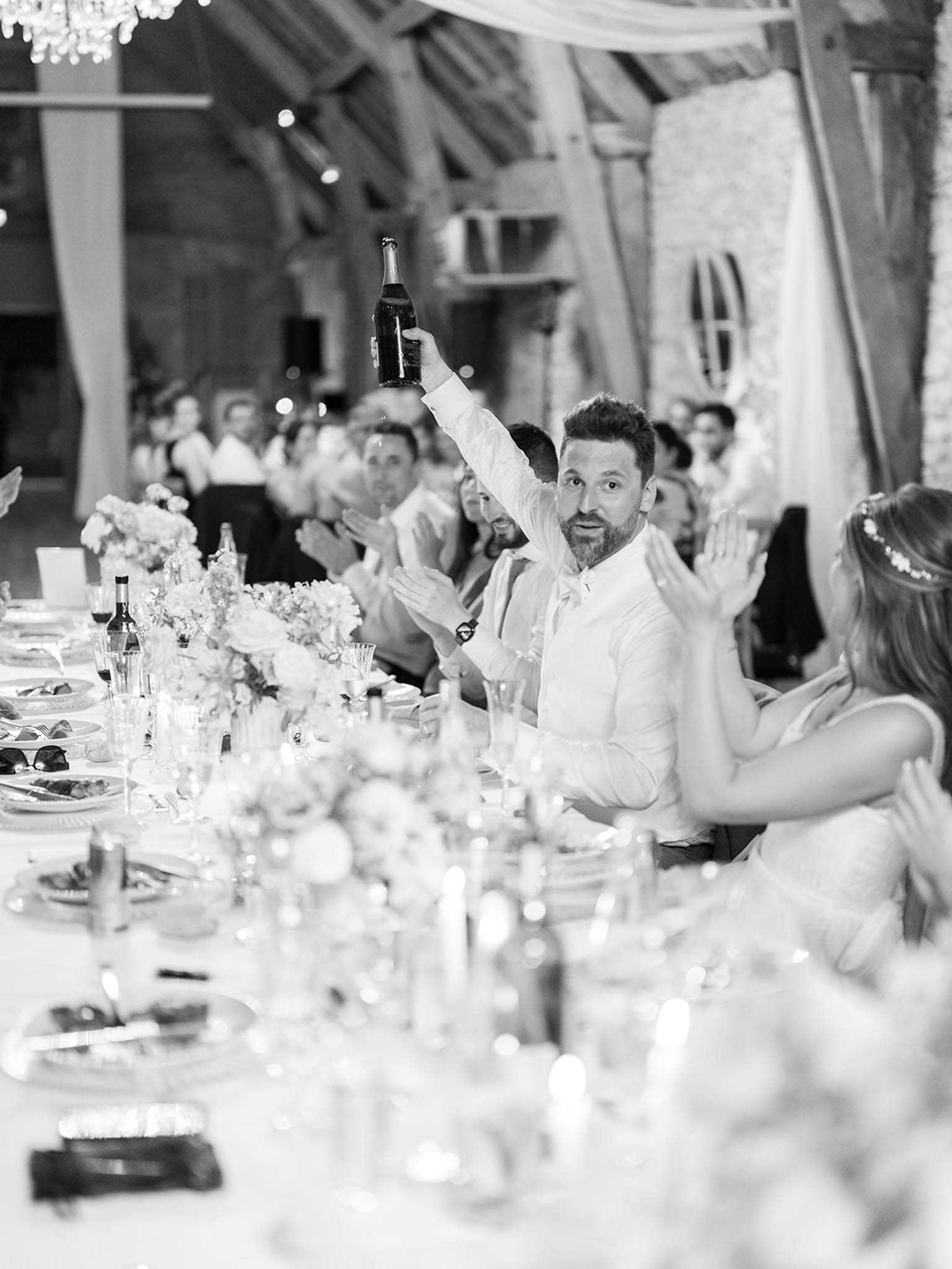 This black-and-white image captures a lively moment during an indoor wedding reception dinner, with a man in a white dress shirt and tie raising a champagne bottle high in the air while guests seated along a long banquet table applaud around him. To his right, a bride wearing a floral hair accessory and a sleeveless gown is clapping, and approximately 15–20 guests are visible in the background doing the same. The venue features exposed wooden beam trusses, draped white fabric, and a chandelier overhead, suggesting a rustic barn or converted agricultural space styled with classic touches. The banquet table is set with glassware, candles, floral centerpieces with rounded blooms and loose arrangements, and scattered place settings; the foreground is softly out of focus, drawing attention to the central figure. The image is a medium wide shot with bright highlights and soft mid-tones typical of film-style photography.