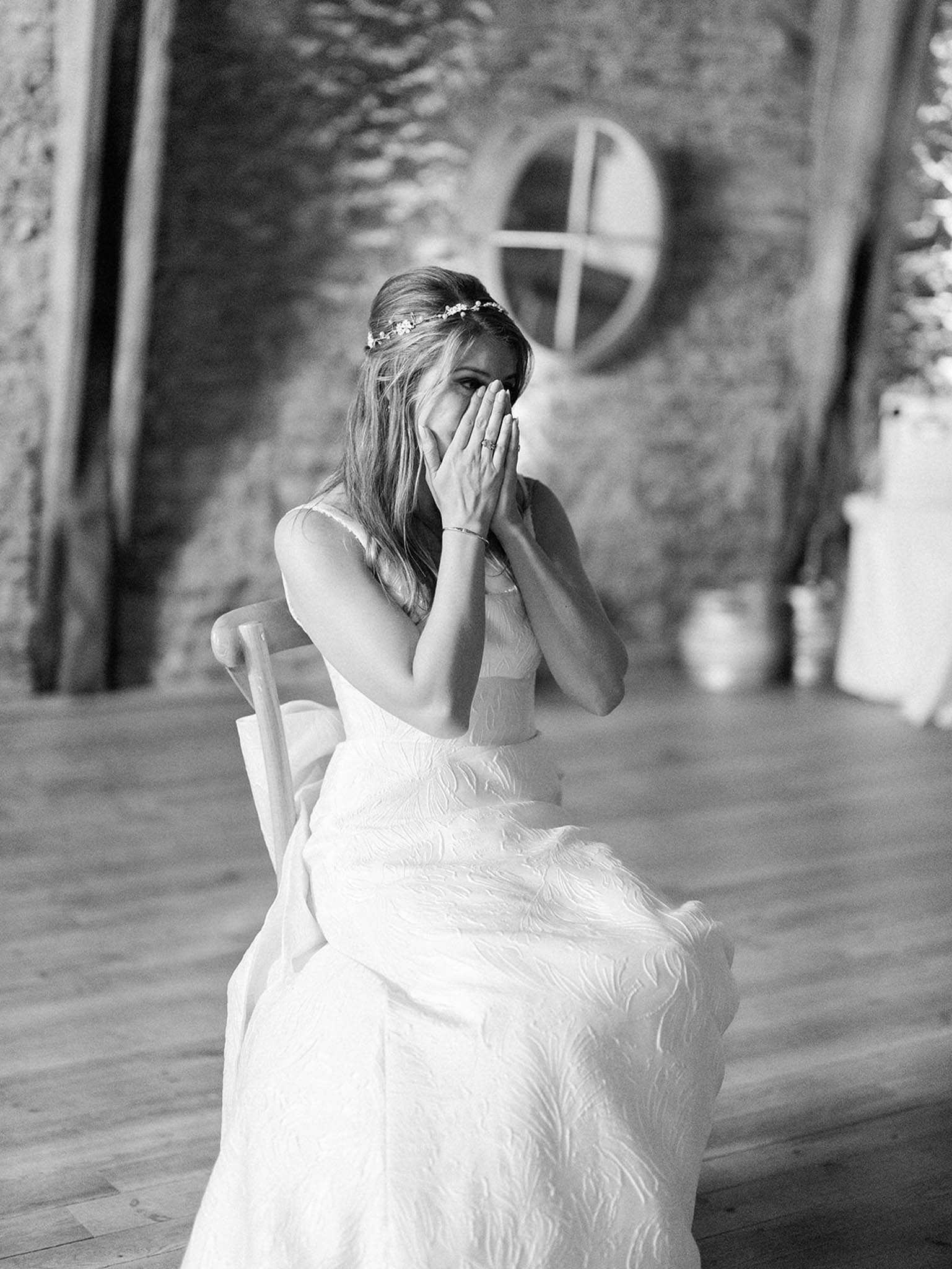 Black-and-white portrait of a bride seated on a wooden chair, covering her face with both hands in an emotional moment, likely during a ceremony or the reading of vows. She wears a textured, embroidered gown with a sleeveless silhouette and a delicate floral hair vine across her half-up hairstyle, with a slim bracelet visible on her wrist. The setting is an indoor rustic stone-walled space with wide-plank wooden floors and a round window visible in the background, suggesting a converted barn or chapel. The image is a medium close-up shot with soft contrast and a shallow depth of field, keeping the background gently out of focus.