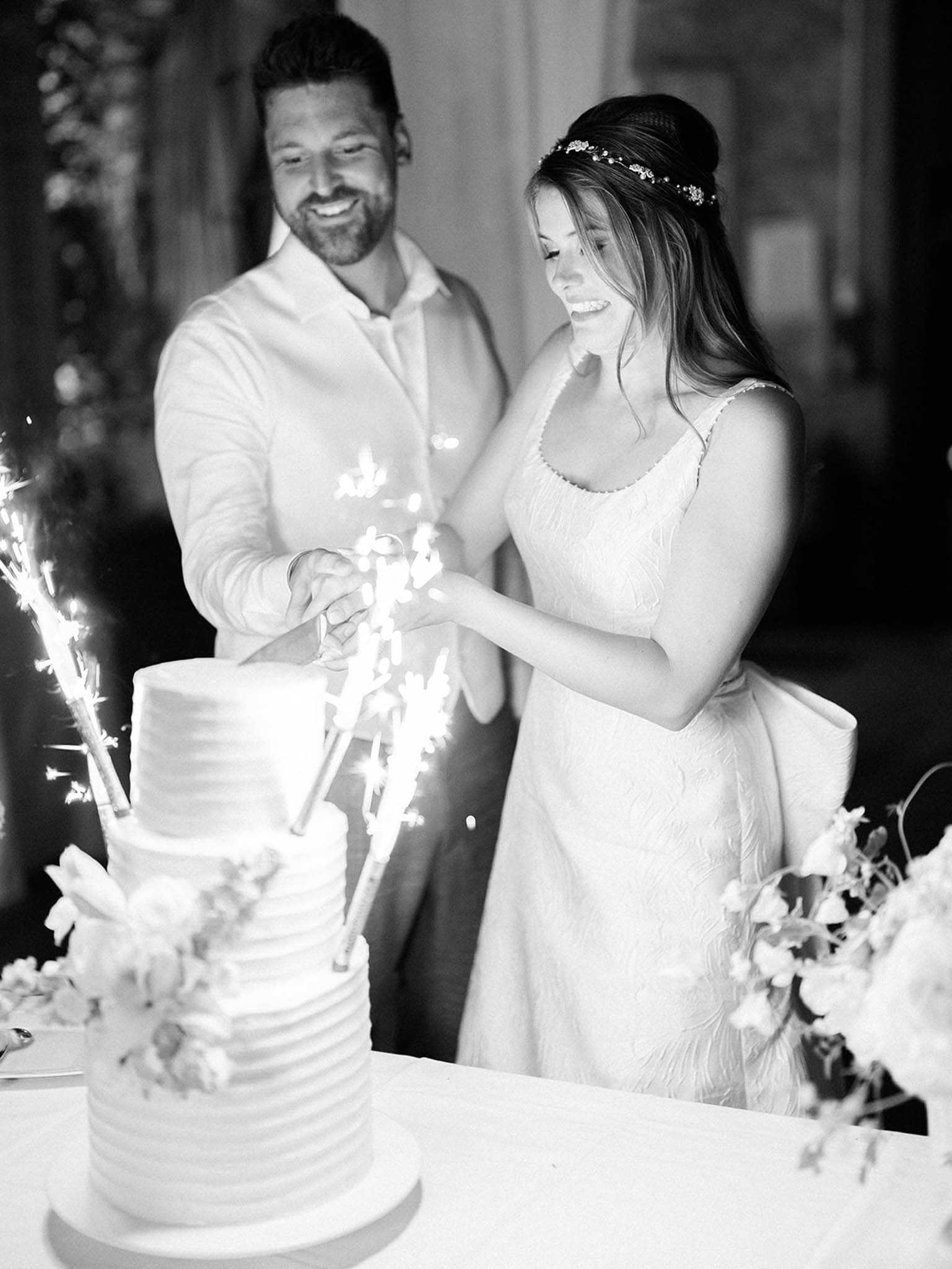 This black-and-white image captures the cake cutting moment at a wedding reception, shot as a mid-range portrait of the couple. The bride wears a textured, form-fitting gown with thin straps and a large back bow, with her hair worn half-up in a bun accented by a delicate floral headpiece; the groom is dressed in a light-colored casual shirt and dark trousers. Both are laughing as they hold a knife together over a four-tier ribbed buttercream wedding cake decorated with small florals at the base, which is topped with lit sparklers creating bright bursts of light against the dark background. A floral arrangement is partially visible in the foreground right, and the setting appears to be an indoor reception space with ornate architectural details softly visible in the background.