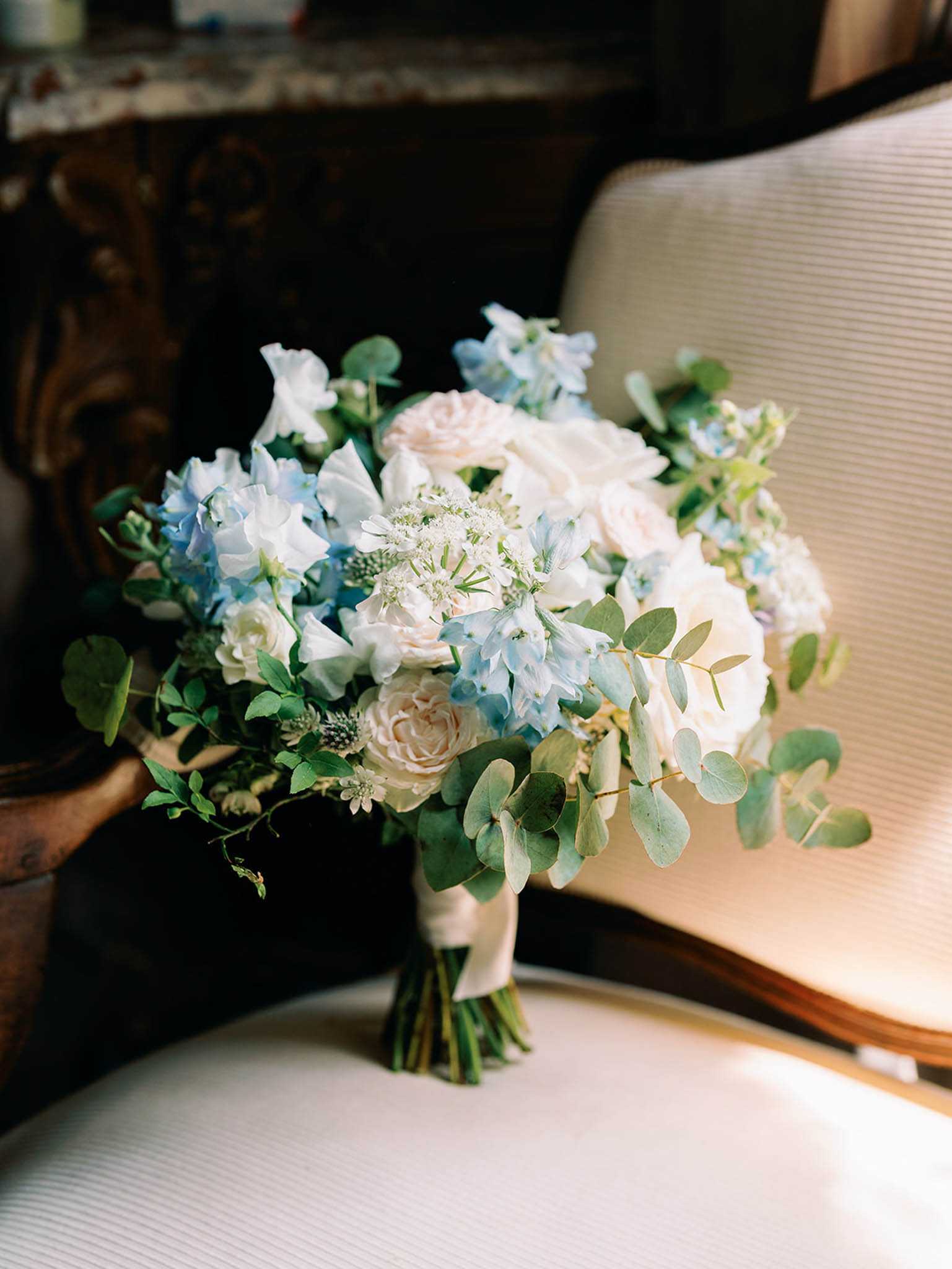 Close-up detail shot of a bridal bouquet resting on a cream upholstered chair, with a dark ornate wooden furniture piece visible in the background. The bouquet is loosely arranged and features pale blush garden roses, powder blue delphinium, white astrantia, white lisianthus, and trailing eucalyptus stems, with small filler greenery throughout. The stems are wrapped in an ivory ribbon. The color palette is soft blue, blush, and white with green accents, suggesting a classic yet garden-inspired floral style.