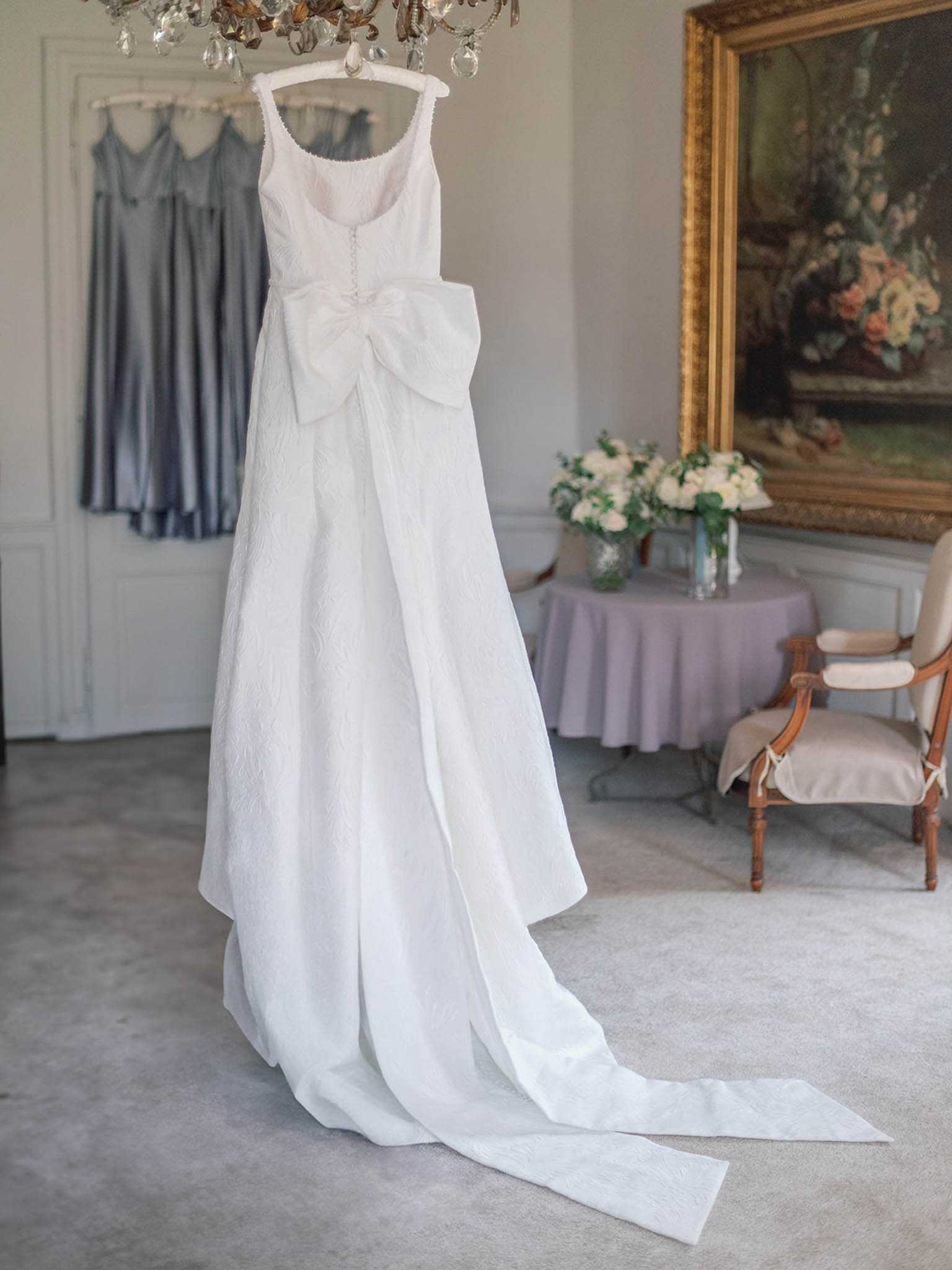 A getting-ready detail shot of a white wedding gown hanging from a crystal chandelier in an ornate interior room. The dress features a scoop neckline, textured jacquard fabric with a subtle floral pattern, a large front bow at the waist, and an extended train pooling on the floor. In the background, two steel-blue satin bridesmaid dresses hang on a door, and a round table covered in a lavender cloth holds a arrangement of white flowers and greenery. A gilded oil painting and a French-style upholstered chair with a wooden frame are visible to the right, suggesting a chateau or historic venue bridal suite. The composition is a full-length portrait shot with soft, even natural lighting.