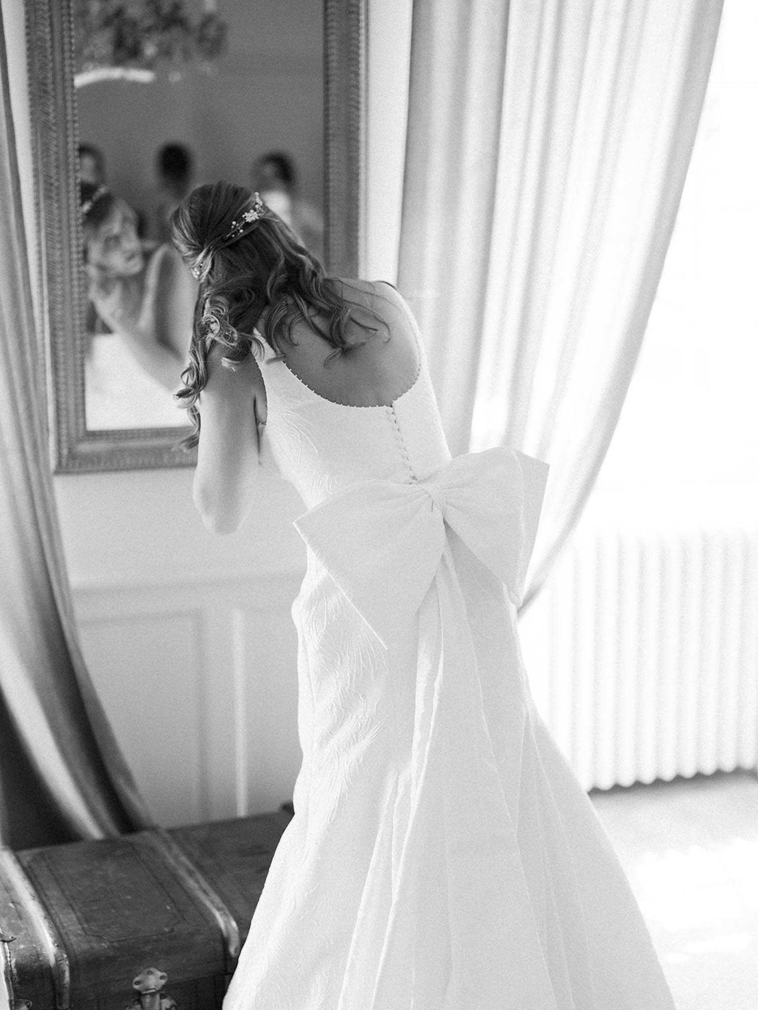 A black-and-white getting-ready portrait of a bride standing with her back to the camera in an indoor room, adjusting her hair or earring with both hands raised. She wears a fitted white gown with a prominent oversized bow at the back waist and a textured fabric with a button-detail spine; her half-up hairstyle is adorned with a delicate hair accessory. The room features paneled walls, floor-length draped curtains with bright backlighting from a large window, and a gilt-framed mirror to the left in which her reflection and the partial reflections of other people in the room are visible. A vintage trunk sits at lower left. The image is a medium portrait shot with soft, high-key tones and gentle contrast.