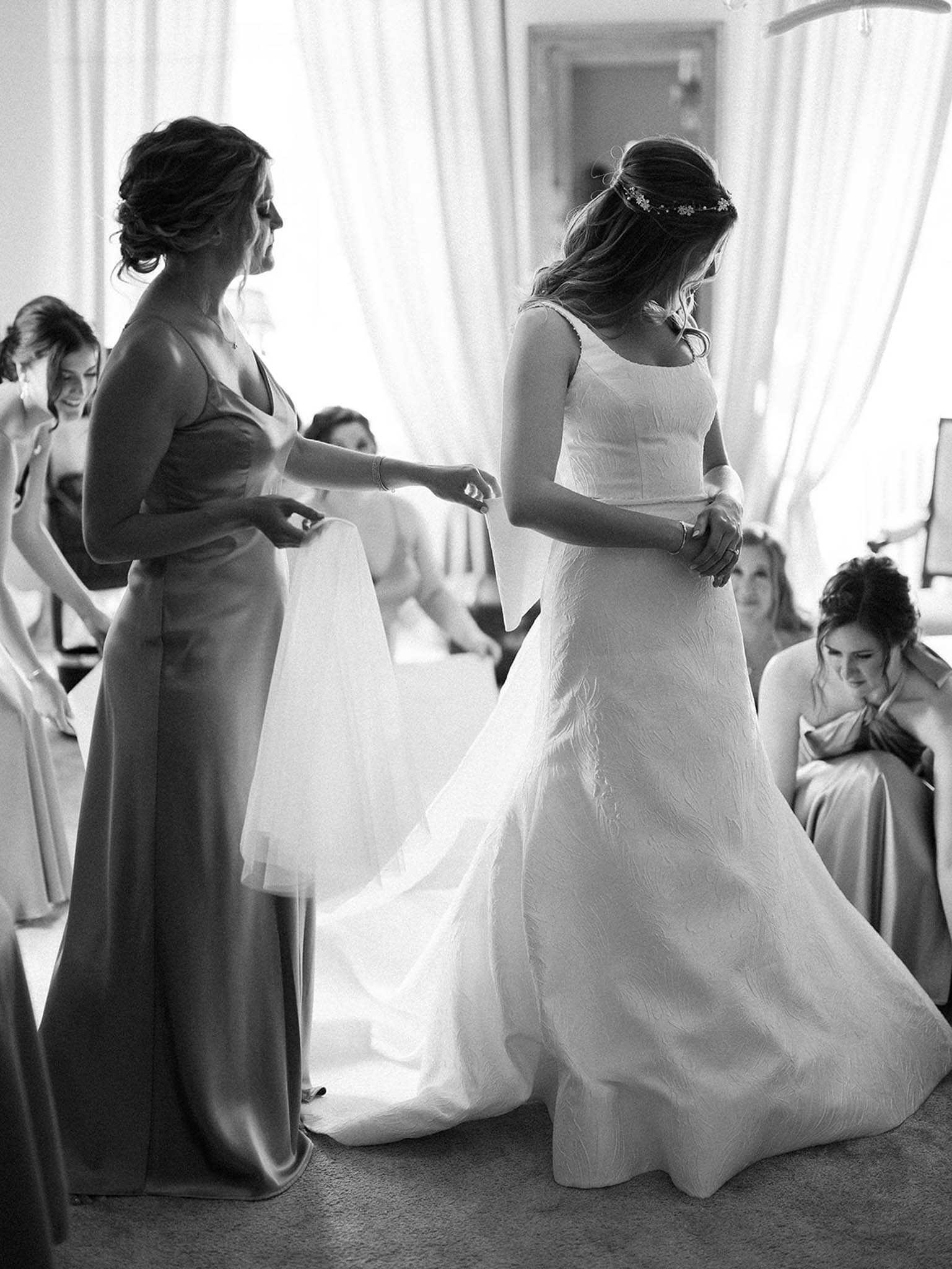 This black-and-white getting-ready scene shows a bride being helped into her veil by a bridesmaid inside what appears to be a bright, curtained room. The bride wears a structured, textured A-line gown with a square neckline and a delicate floral headpiece, her hair worn half-up. The bridesmaid facing her wears a floor-length satin slip-style dress with a V-neckline. In the background, approximately three additional bridesmaids in matching satin gowns are visible — some seated and adjusting their own attire. The image is shot in medium contrast black and white, with bright backlighting from sheer curtains creating soft highlights around the figures. The composition is a mid-range portrait shot capturing the interaction between the bride and bridesmaid at the center of the frame.