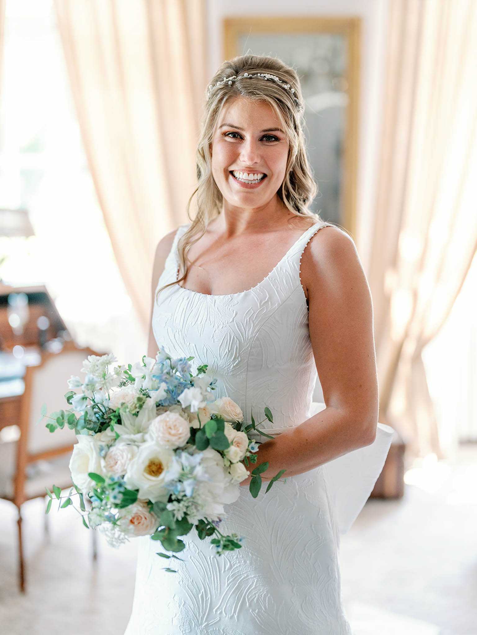 A bridal portrait taken indoors in a well-lit room with sheer cream curtains and a gold-framed mirror visible in the background. The bride wears a fitted white sleeveless gown with an embossed floral texture and a scoop neckline, accessorized with a delicate pearl and crystal hair vine worn across a half-up hairstyle. She holds a lush, loosely arranged bouquet featuring blush garden roses, white ranunculus, pale blue delphinium, white sweet peas, and trailing eucalyptus. The composition is a mid-length portrait shot with soft natural backlit lighting.