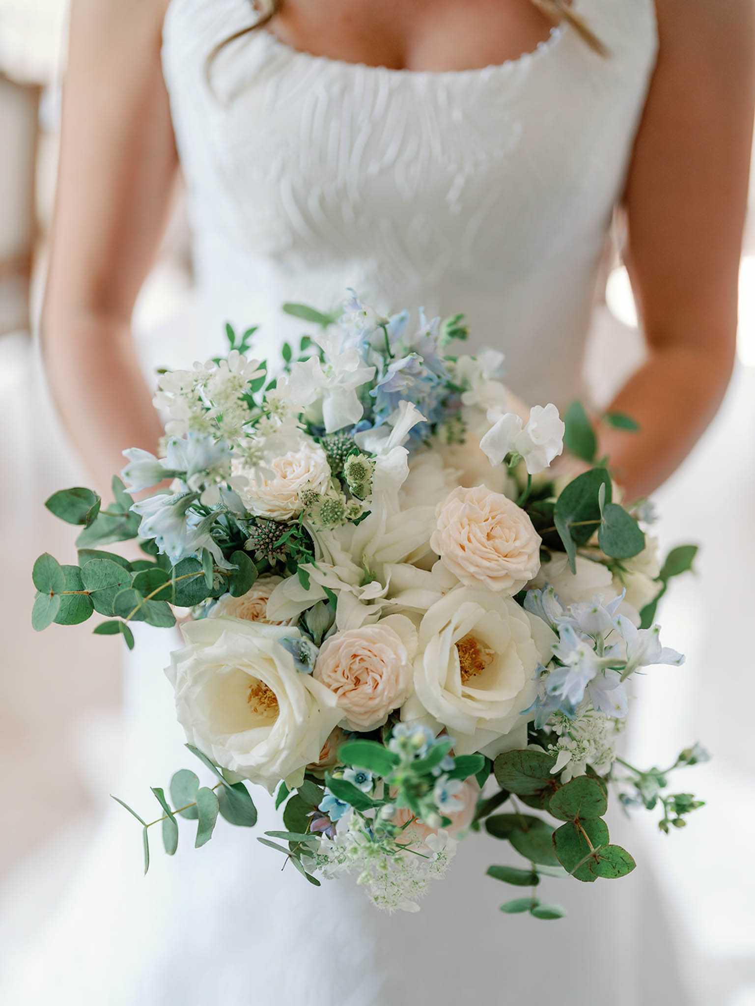 A close-up detail shot of a bride holding her bridal bouquet, with the bride's torso visible in the background wearing a white ruched strapless gown. The bouquet is a loosely arranged, garden-style composition featuring ivory garden roses, blush peach spray roses, pale blue delphinium, white sweet peas, white astrantia, and abundant eucalyptus foliage throughout. The color palette is soft and muted, combining ivory, blush, and powder blue tones against lush green eucalyptus. The bouquet is full and rounded with trailing foliage giving it an organic, unstructured feel.