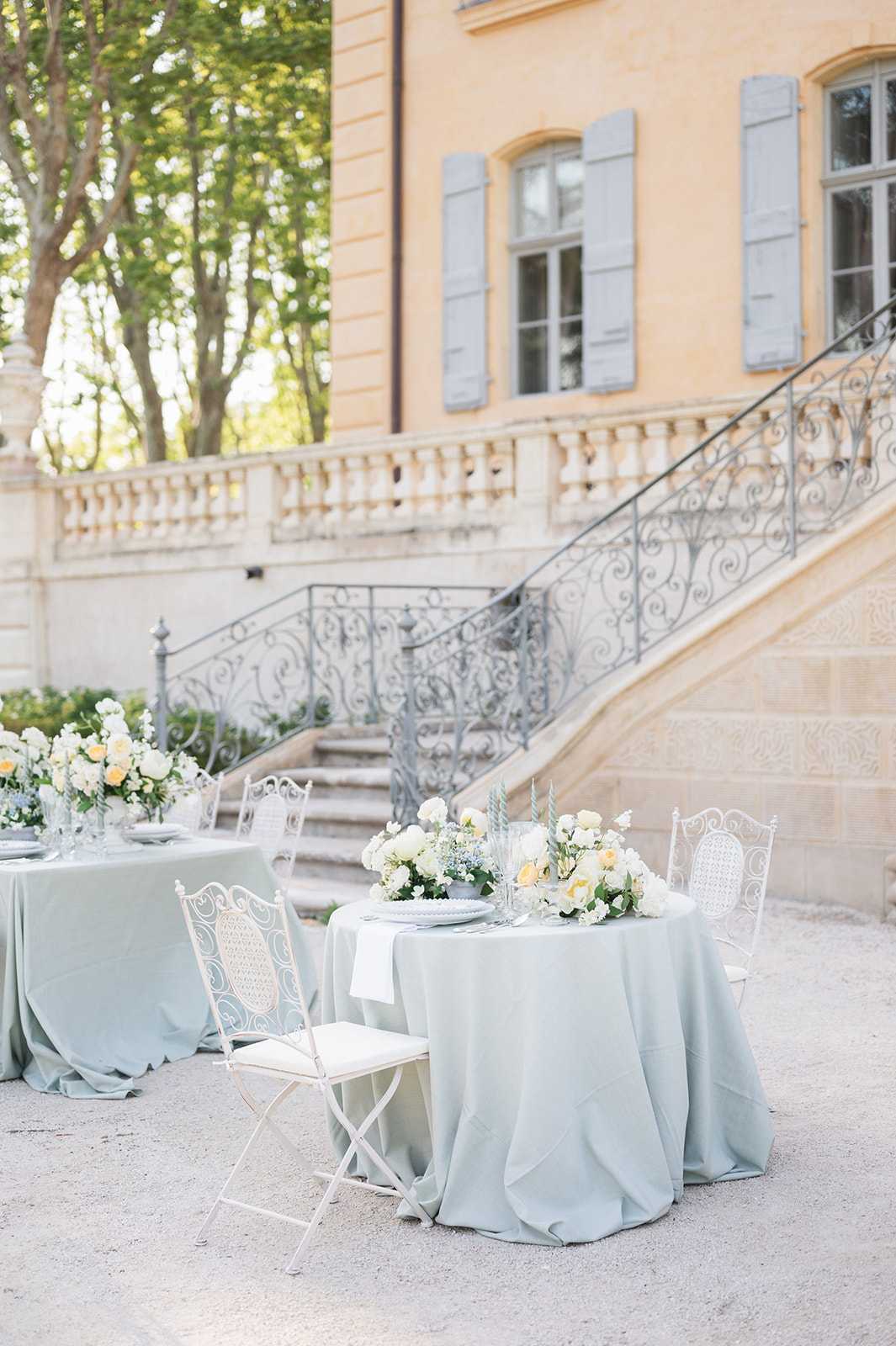 An outdoor wedding reception tablescape set up on a gravel courtyard in front of a French chateau with warm ochre-toned stone walls, grey-blue shutters, and ornate wrought-iron staircase railings with scrollwork detail. Two round tables are dressed in floor-length sage green linen tablecloths, surrounded by white ornate iron bistro-style folding chairs with lattice backs. The foreground table is topped with a lush floral centerpiece of ivory roses, soft yellow roses, white blooms, and touches of light blue flowers with greenery, alongside twisted blue-green taper candles and white stacked plates. A second similarly styled table is visible to the left. The overall decor palette is sage green, ivory, soft yellow, and pale blue, giving a classic French garden aesthetic. Wide shot capturing both the table styling and the chateau facade as backdrop. Potential venue feature image.