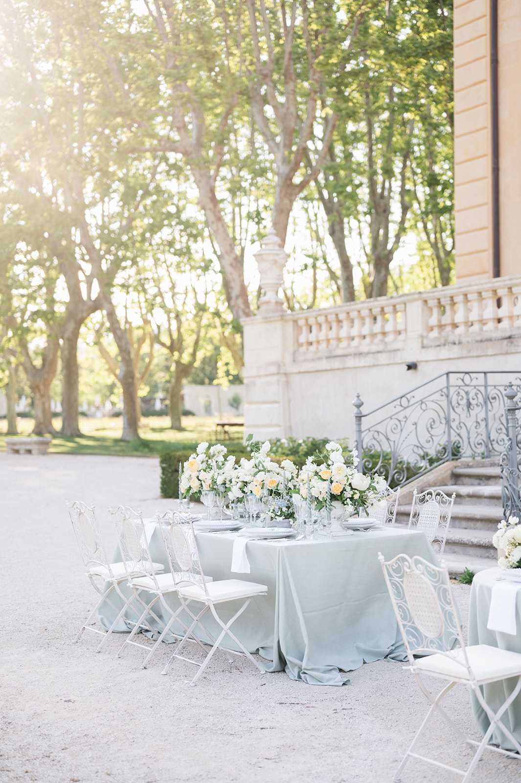 An outdoor wedding reception tablescape set on a gravel forecourt beside a classical French chateau facade featuring a stone balustrade and ornate wrought-iron staircase railing. The rectangular table is dressed in a pale sage-blue linen tablecloth and surrounded by white vintage-style wrought-iron folding chairs with white cushioned seats. The centerpiece consists of a lush low arrangement of ivory and soft peach garden roses with greenery, placed in clear glass vessels alongside glassware and white place settings. The styling is classic French with a soft, muted color palette of sage, white, and ivory. Wide establishing shot taken in warm afternoon light with no people present. Potential venue feature image.