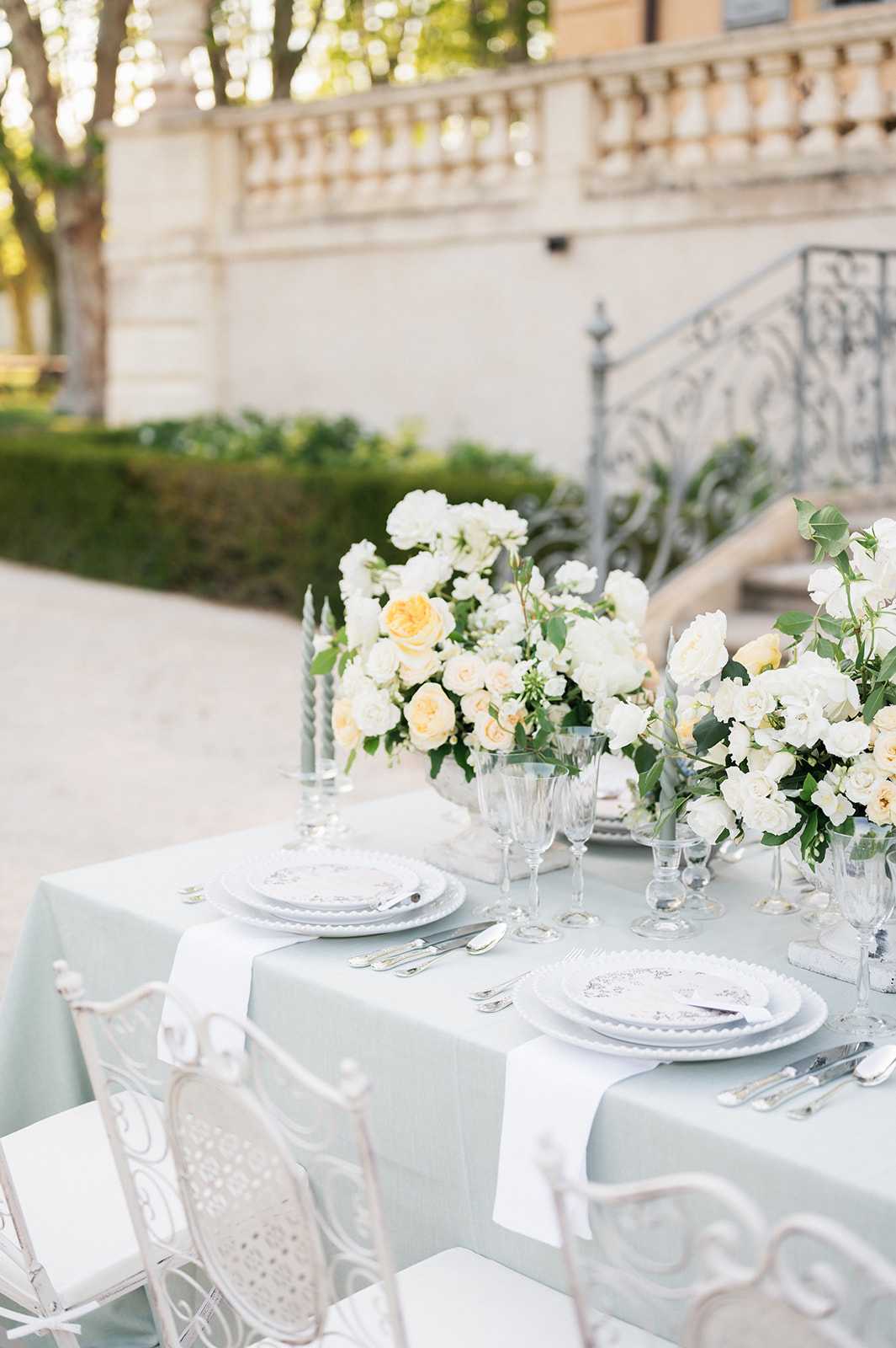 A close-up detail shot of an outdoor wedding reception tablescape set on a terrace or courtyard in front of a classical stone balustrade with ornate wrought-iron staircase railings. The table is dressed in a soft sage-green linen with white linen napkins and set with white scallop-edged plates stacked on grey chargers, silver flatware, and clear crystal glassware. Two lush floral centerpieces feature white garden roses, cream and soft yellow David Austin roses, white lisianthus, and greenery, flanked by slim silver taper candles. White ornate wrought-iron chairs surround the table, consistent with a classic French garden aesthetic. No people are present in the image.