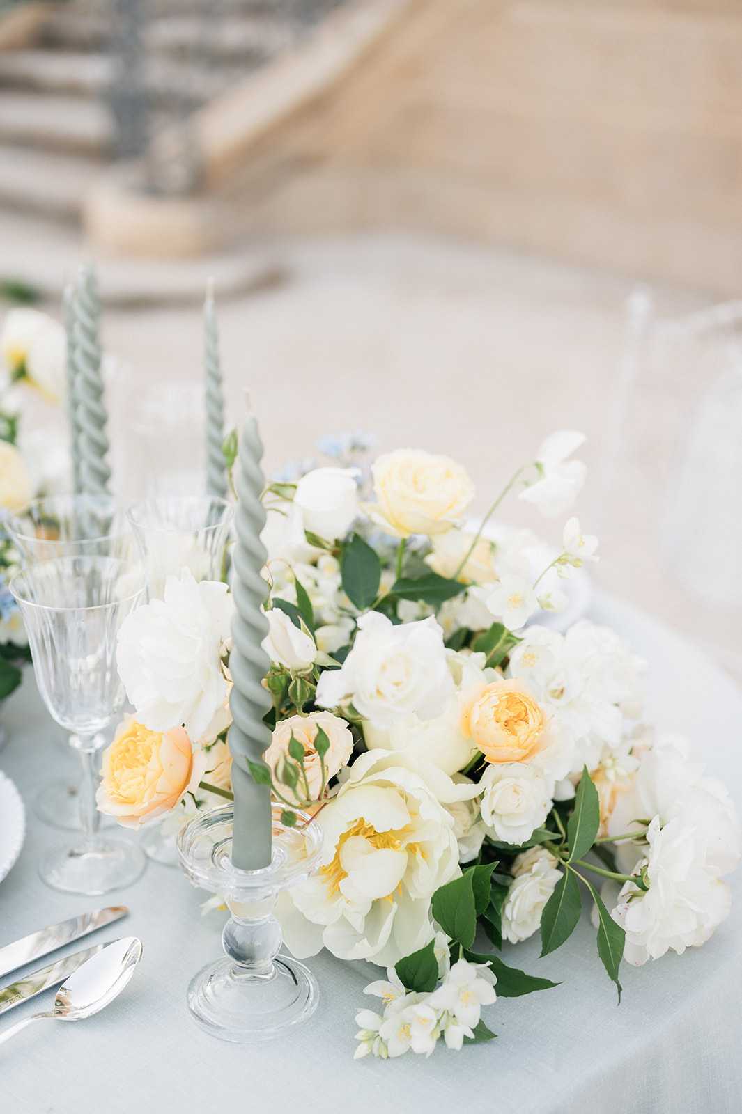 Close-up detail shot of an outdoor wedding reception table setting featuring a low, lush floral centerpiece composed of ivory peonies, peach garden roses, white spray roses, small white jasmine-like blooms, and touches of pale blue delicate flowers with green foliage. Two sage green twisted taper candles in clear glass candlestick holders are placed within the arrangement. The table is dressed with a pale blue-grey linen, and crystal etched glassware alongside silver flatware are visible at the place setting. The overall decor palette is soft and airy, combining ivory, peach, sage green, and pale blue in a classic French romantic style.