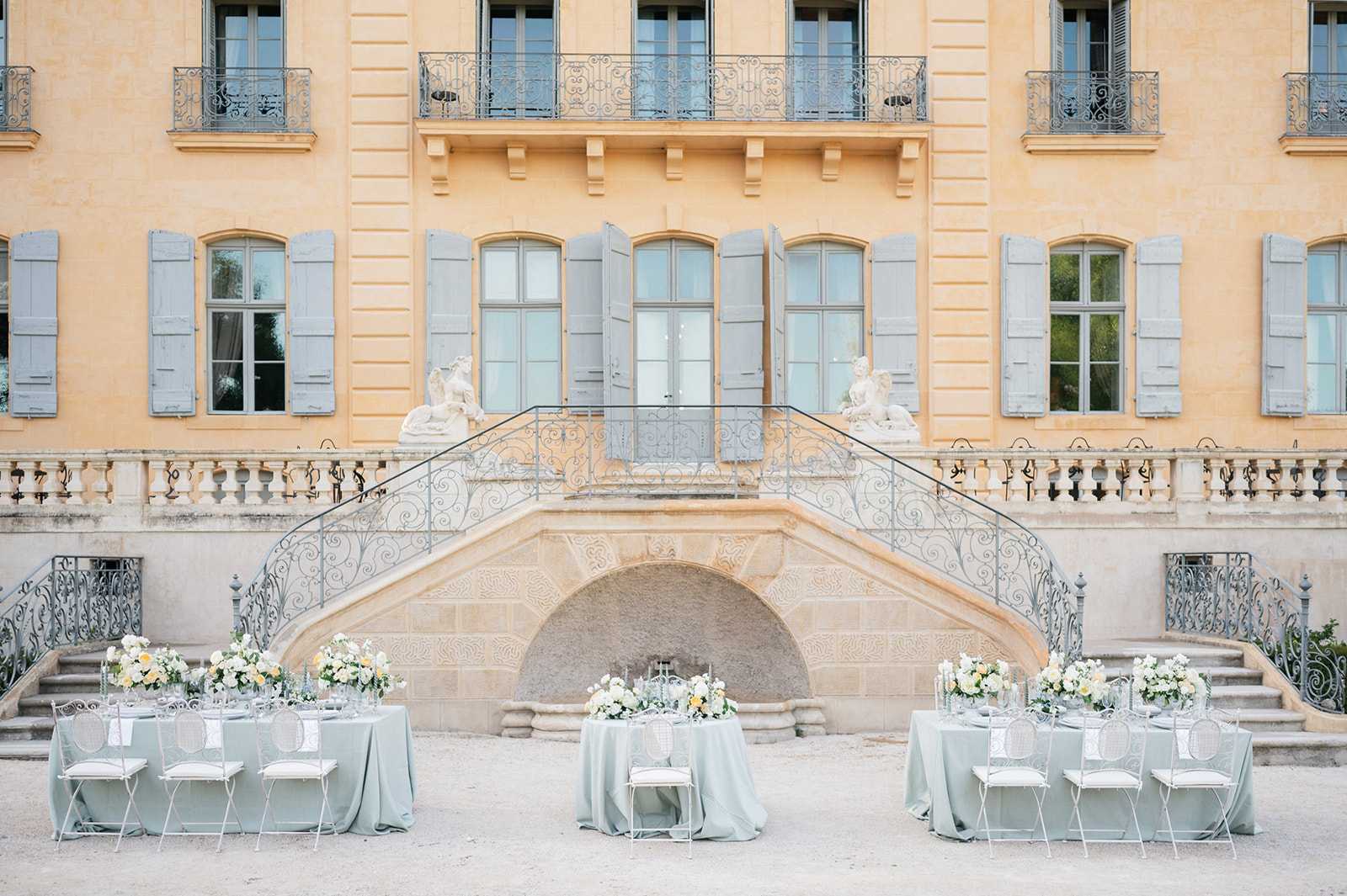 Outdoor reception setup in the courtyard of a French chateau, with three round tables dressed in sage green floor-length linen tablecloths arranged symmetrically in front of the venue's grand double staircase. The wrought-iron staircase features ornate scrollwork and is flanked by two white stone figurative sculptures, with the chateau facade displaying warm ochre-toned render, grey-blue shuttered arched windows, and iron-railed balconies above. Each table is styled with low floral centerpieces composed of ivory, white, and soft yellow blooms — likely roses, ranunculus, and similar garden flowers — alongside taper candles and glassware. The chairs are white metal with a medallion back design and white seat cushions, consistent with a classic French aesthetic. The wide shot captures the full architectural facade as the primary backdrop, with no people present. Potential venue feature image.