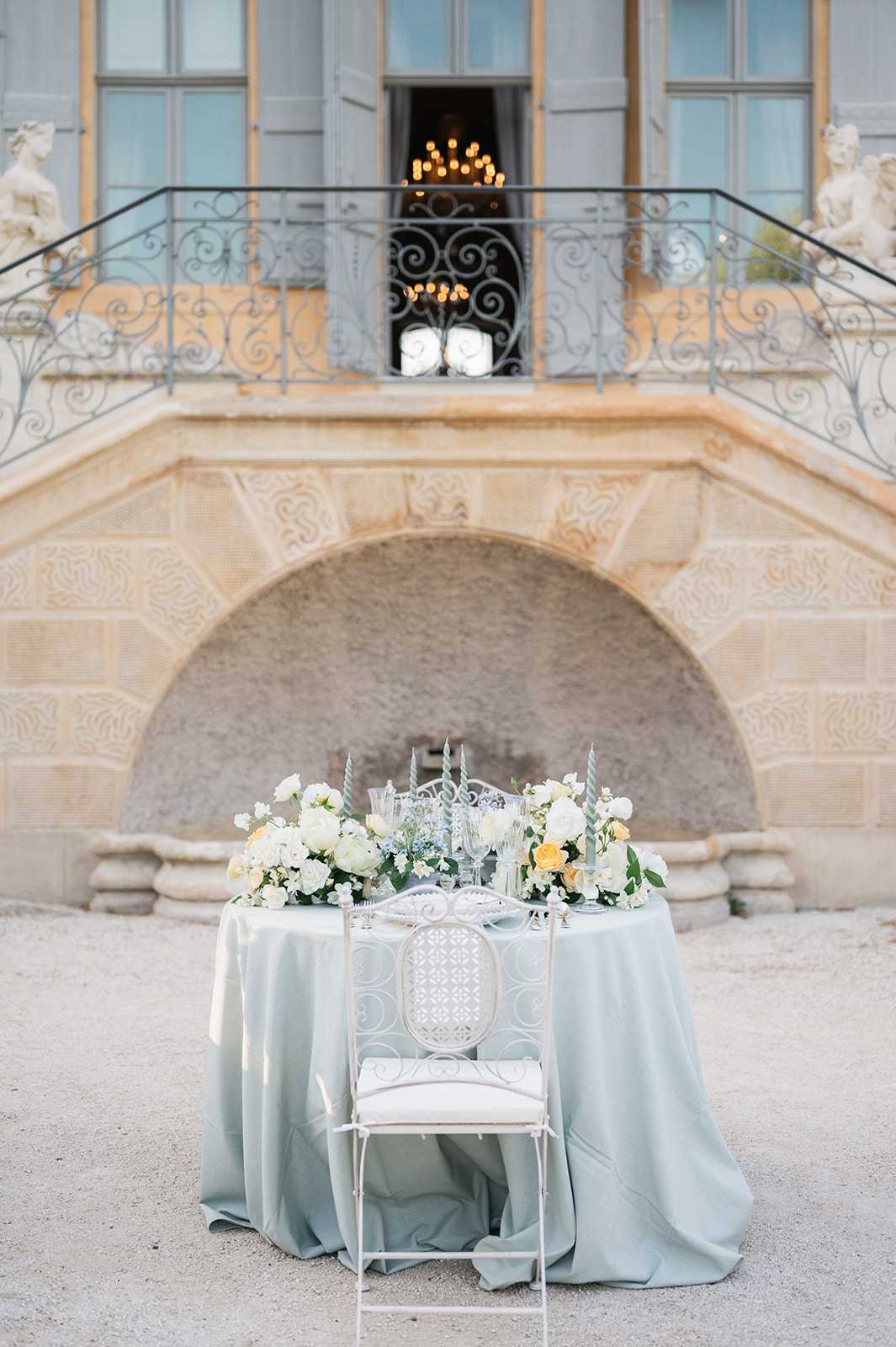 A styled sweetheart table setup photographed outdoors in the gravel courtyard of a French chateau, with no people present. The small round table is draped in a sage green silk tablecloth that pools on the ground, topped with low floral arrangements of ivory garden roses, soft yellow roses, white ranunculus, and delicate filler blooms in white and pale blue. Crystal glassware, white place settings, and pale grey twisted taper candles are arranged at the center. A single ornate white wrought-iron chair with a cream cushion faces the camera. The backdrop features the carved limestone facade of the chateau, including an arched niche, decorative stone reliefs, sculptural figures flanking a wrought-iron balustrade staircase, and an open window through which a glowing chandelier is visible. The overall decor palette is sage green, ivory, and soft yellow with a classic French formal styling. Wide shot, slightly low angle. Potential venue feature image.