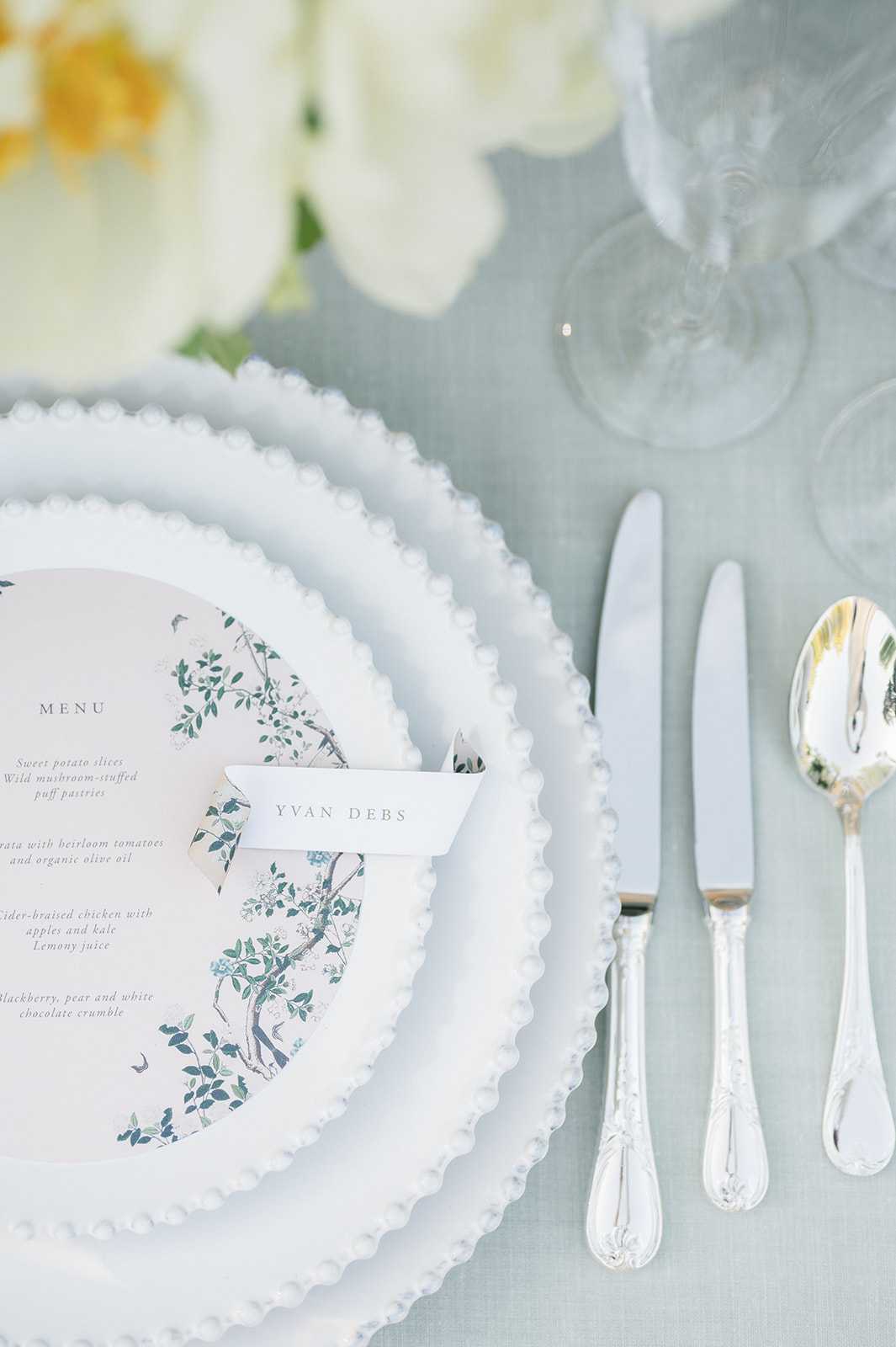 A close-up detail shot of a wedding reception place setting on a pale blue-grey linen tablecloth. The place setting features stacked white beaded-edge charger and dinner plates, topped with a blush pink circular menu card printed with a chinoiserie botanical motif in teal and green, alongside a small white place card reading 'YVAN DEBS'. Silver ornate-handled cutlery — two knives and a spoon — is arranged to the right, and two crystal wine glasses are visible in the upper right corner. White flowers, likely peonies, are softly blurred in the upper left background. The overall palette is soft blue-grey, white, blush, and silver, consistent with a classic French or European-style reception tablescape.