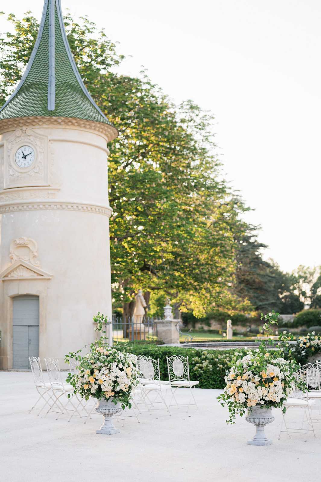 An outdoor wedding ceremony setup on a gravel terrace beside a French chateau pavilion featuring ornate cream stone architecture, decorative carved moldings, a clock face, and a green tiled conical turret roof. Two large white stone urns flank the aisle, each filled with full arrangements of cream and soft peach roses, white blooms, and trailing green ivy and foliage. White wrought-iron bistro-style chairs are arranged in rows on either side of the aisle, with a small white ornate table visible at the ceremony focal point. The overall decor palette is white, cream, and soft peach with green accents, consistent with a classic French garden aesthetic. Wide shot capturing both the architectural setting and the ceremony layout. Potential venue feature image.