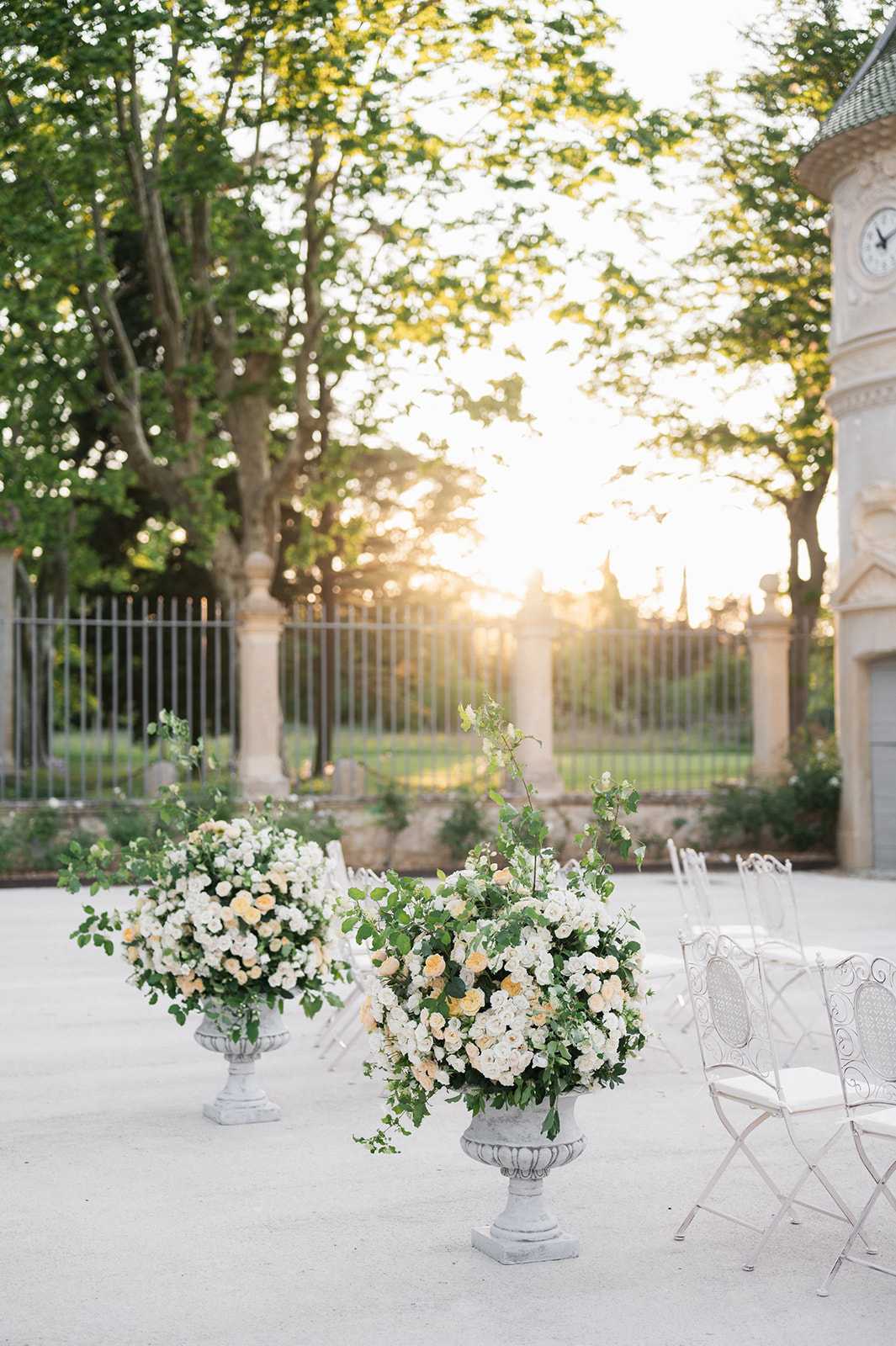 An outdoor ceremony setup at what appears to be a French chateau, photographed at golden hour with warm backlight filtering through the gates and trees. Two large stone urn pedestals hold abundant floral arrangements composed of ivory and soft peach garden roses, white spray roses, and trailing green foliage with climbing vine stems. White wrought-iron folding chairs are arranged in rows to the right, and a classical stone pavilion with a clock tower is partially visible on the right edge of the frame. The overall decor palette is white, cream, and soft peach with lush greenery, consistent with a classic French garden ceremony aesthetic. Wide shot taken from ground level, emphasizing the floral urns in the foreground against the backlit courtyard setting. Potential venue feature image.