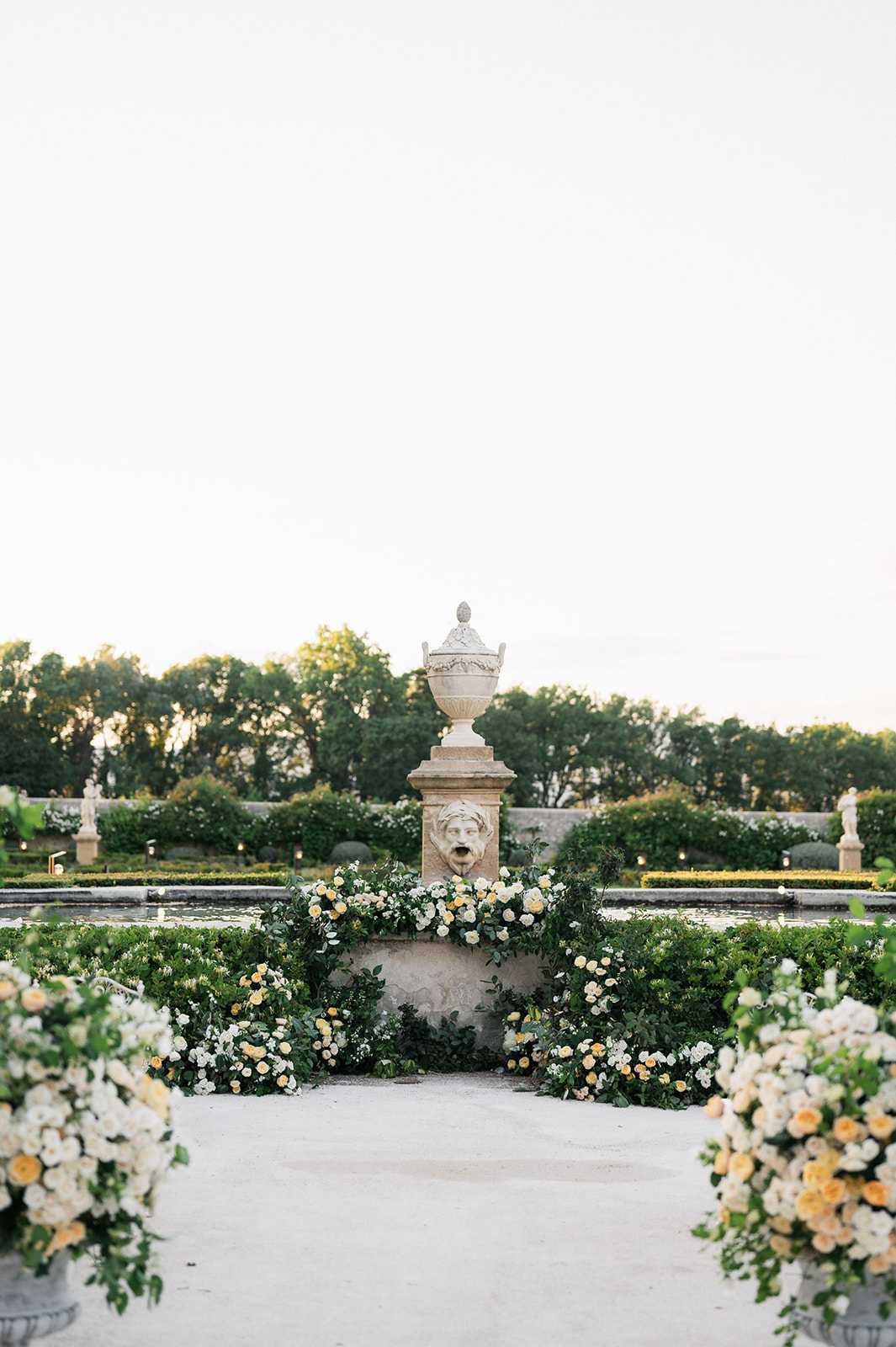 An outdoor ceremony or cocktail hour setup in a formal French garden, centered on a classical stone fountain pedestal with a carved face mask and ornamental urn on top. The base of the pedestal is surrounded by a dense ground-level floral installation of ivory and soft peach-yellow roses with trailing greenery. In the foreground on either side of a gravel aisle, large arrangement urns hold full clusters of ivory and peach-yellow roses with green foliage. The garden beyond features neatly clipped boxwood hedges, a reflecting pool, and stone statuary, styled in a classic formal French garden aesthetic. The composition is a wide symmetrical shot taken at ground level along the central axis of the garden. Potential venue feature image.