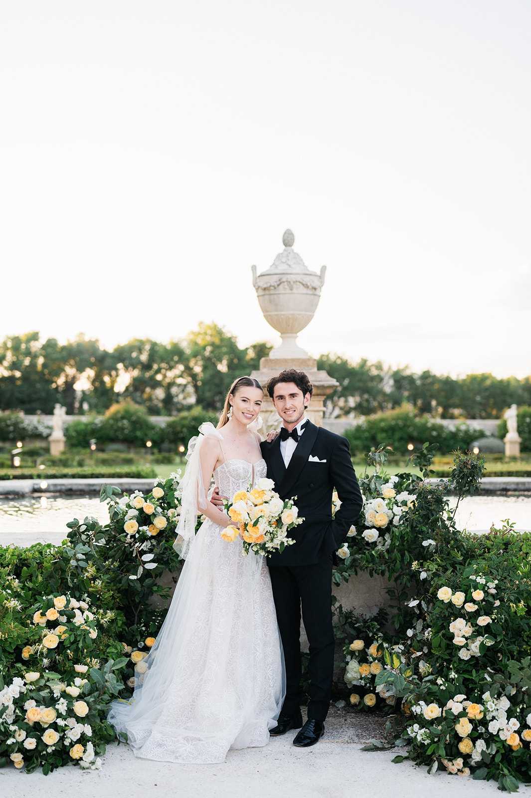 A couple poses together for a portrait in a formal French garden setting, standing in front of a large ornamental stone urn on a pedestal flanked by a reflecting pool and classical statues in the background. The bride wears a white lace ballgown with a corset bodice, spaghetti straps, a tulle skirt, and a cathedral-length veil with a bow detail at the shoulder; she carries a lush bouquet of yellow and cream garden roses with white ranunculus and greenery. The groom wears a black double-breasted tuxedo with a white pocket square and black bow tie. The couple is surrounded at ground level by an abundant low floral arrangement of yellow and white garden roses and lush green foliage, extending across the full width of the frame in a classic, formal style. The shot is a mid-length portrait taken at golden hour.