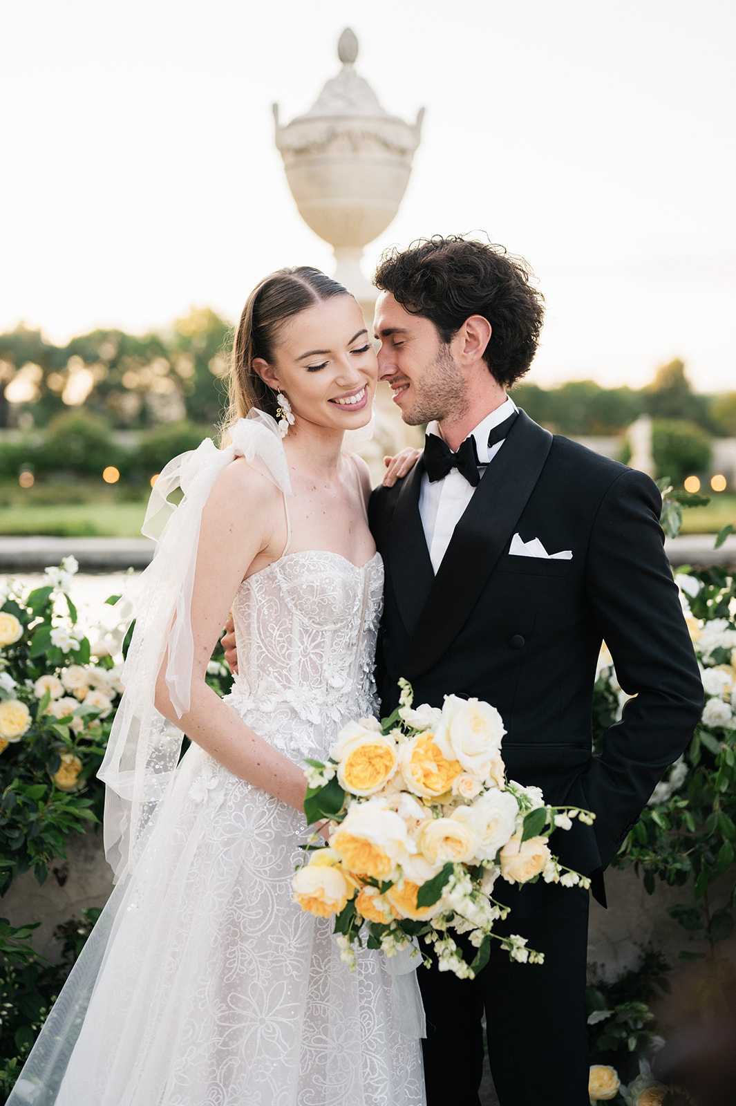 A couple portrait taken outdoors at golden hour on what appears to be a formal garden terrace, with a large stone urn pedestal visible in the background. The bride wears a strapless, heavily embroidered and beaded ball gown with floral lace detailing, an organza bow at the shoulder, and drop earrings; she holds a loose bouquet of yellow garden roses, cream peonies, and white sweet peas with trailing greenery. The groom wears a black tuxedo with peak lapels, black bow tie, and white pocket square. Behind the couple is a low stone balustrade decorated with a lush garland of yellow and cream roses, white blooms, and deep green foliage, consistent with a classic formal French garden setting. The composition is a mid-length portrait shot with soft bokeh in the background.